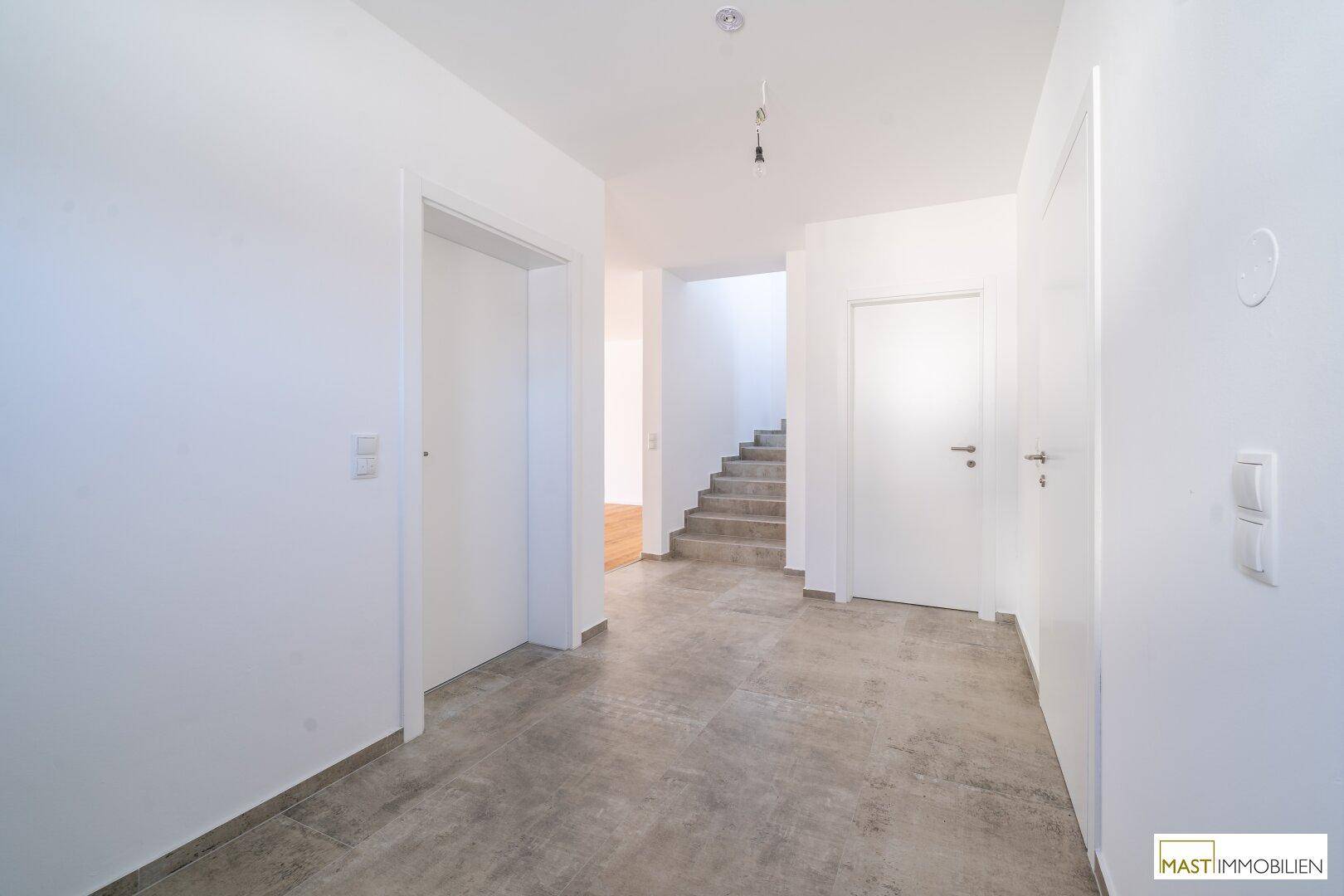 Geräumiger Flur mit Fliesenboden, weißen Wänden und Blick auf die Treppe ins Obergeschoss.
