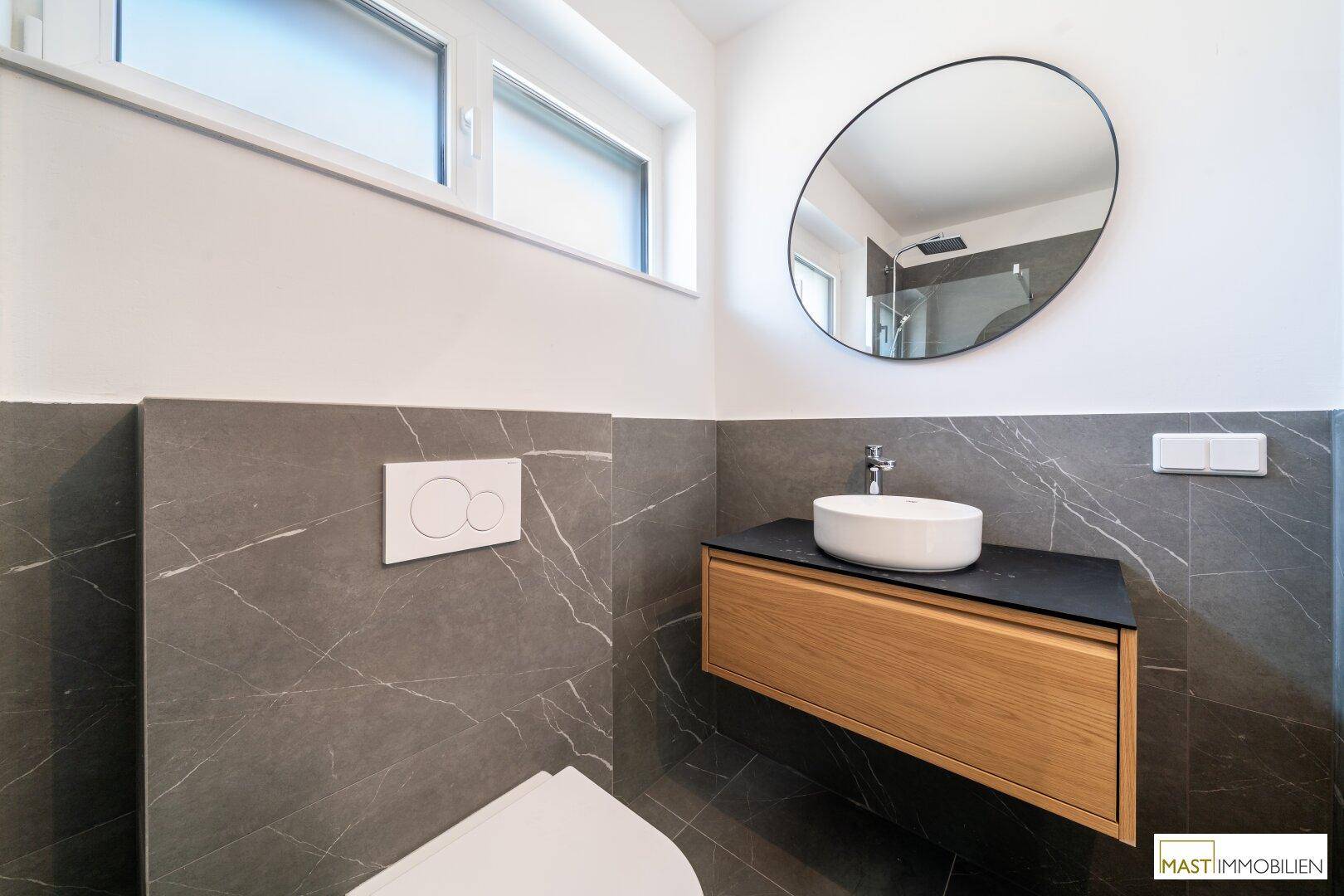 Modernes Badezimmer mit dunklen Marmorfliesen, schwebendem Waschtisch und rundem Spiegel.