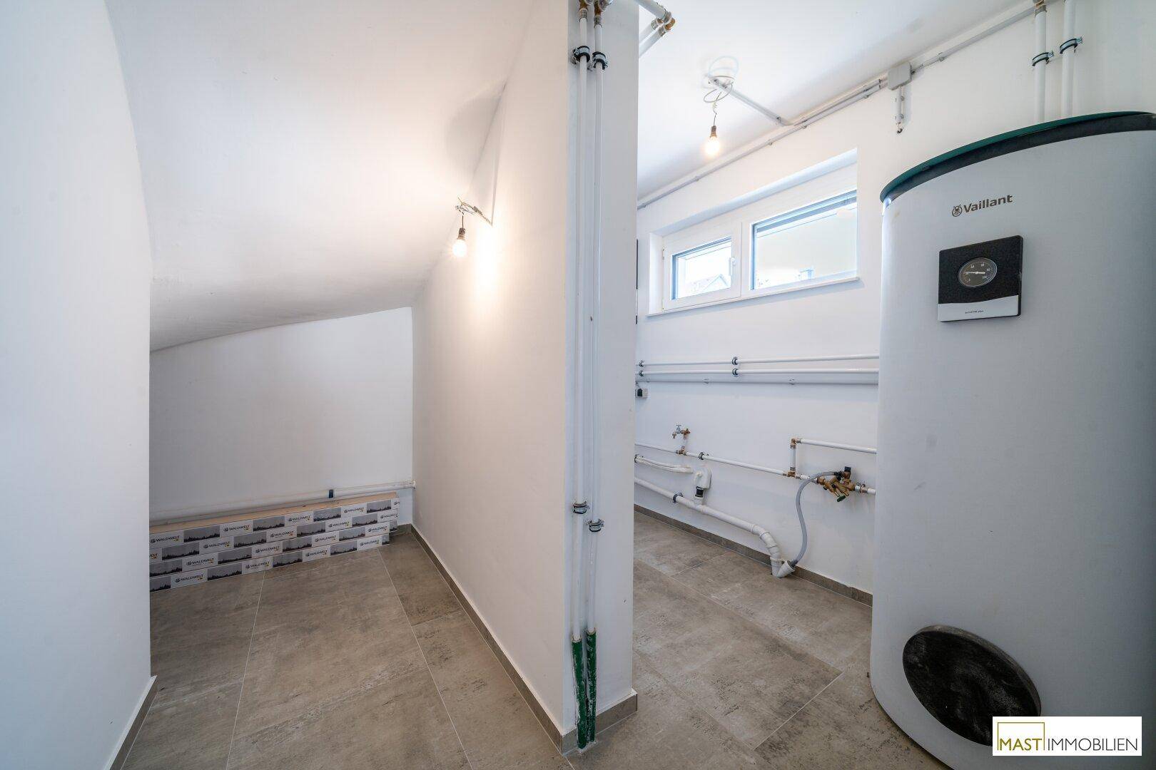 Technikraum mit Warmwasserspeicher, sichtbaren Leitungen und Fenster für ausreichende Belüftung.