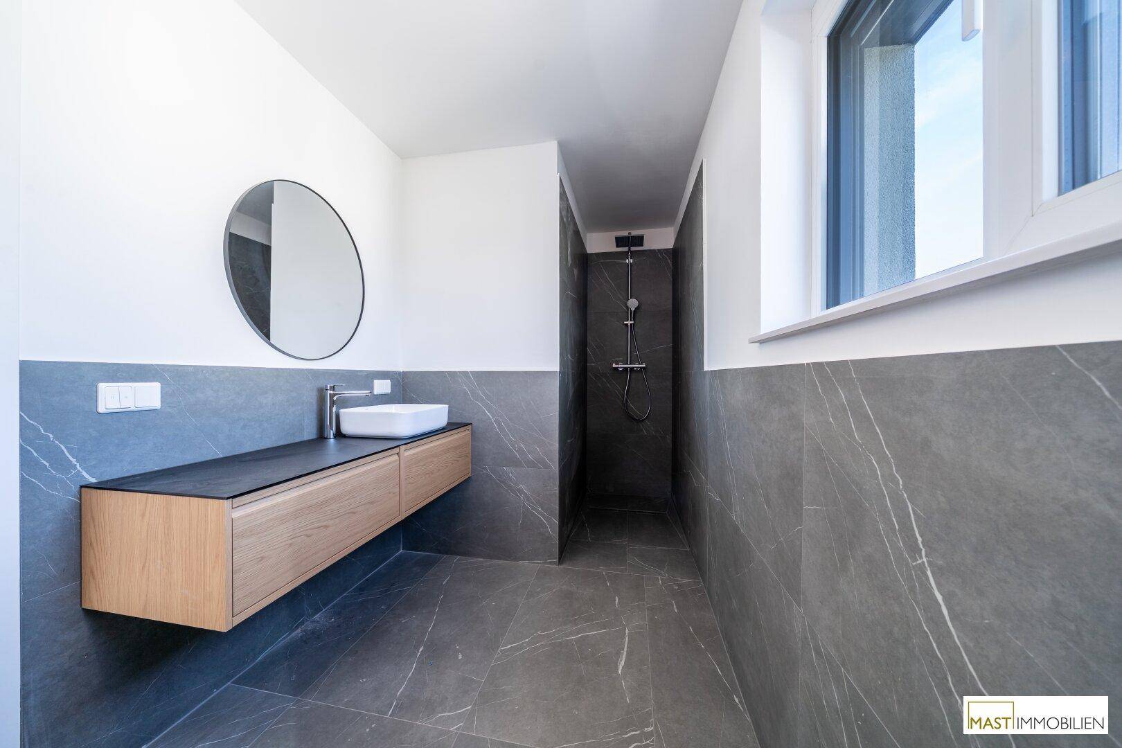 Geräumiges Badezimmer mit moderner Dusche, schwebendem Waschtisch und viel Tageslicht durch das Fenster.