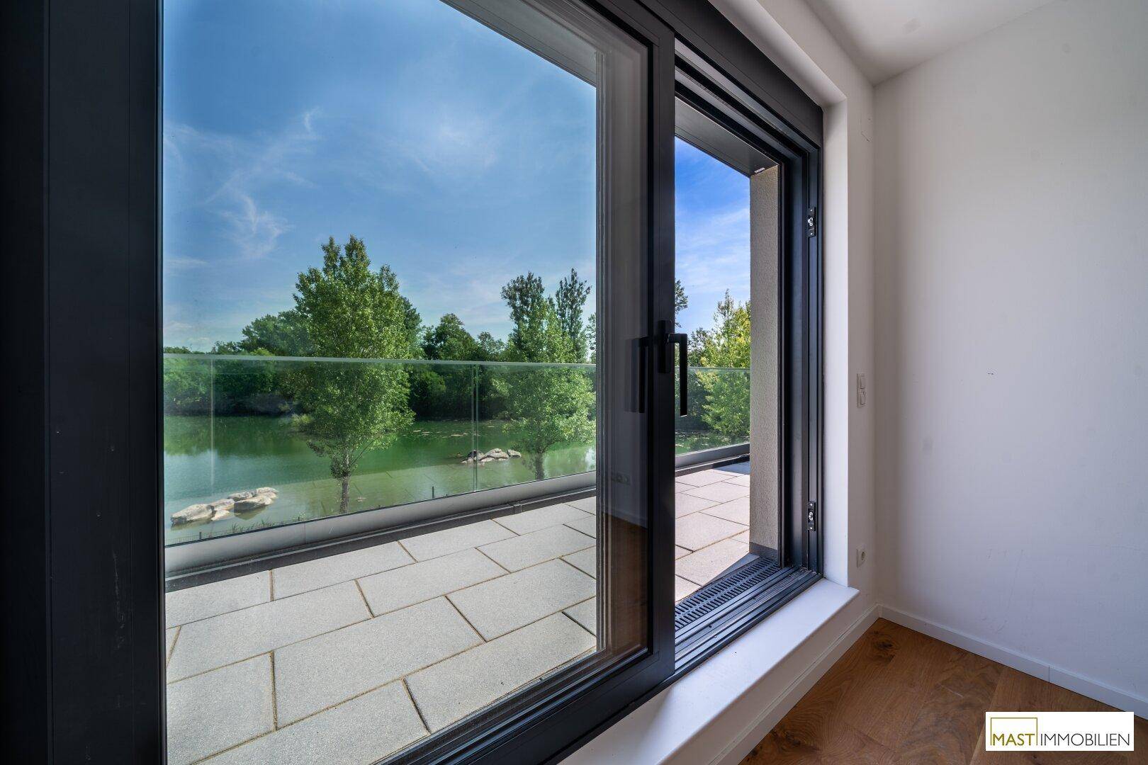 Innenansicht mit großem Schiebefenster, das den Zugang zum Balkon und den Blick auf den See freigibt.