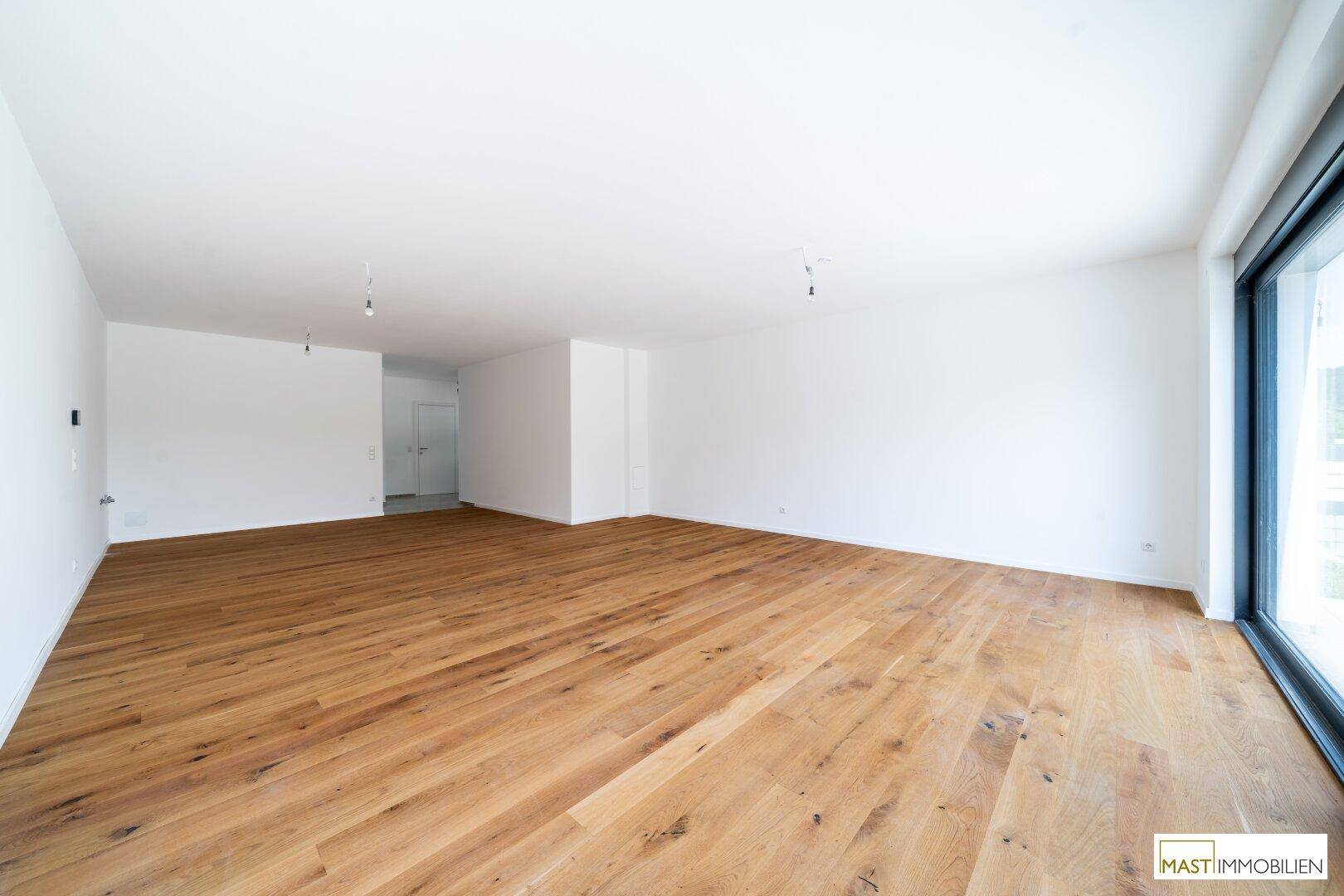 Weitläufiger Wohnbereich mit hellem Holzfußboden und großen Fenstern, die viel Tageslicht hereinlassen.