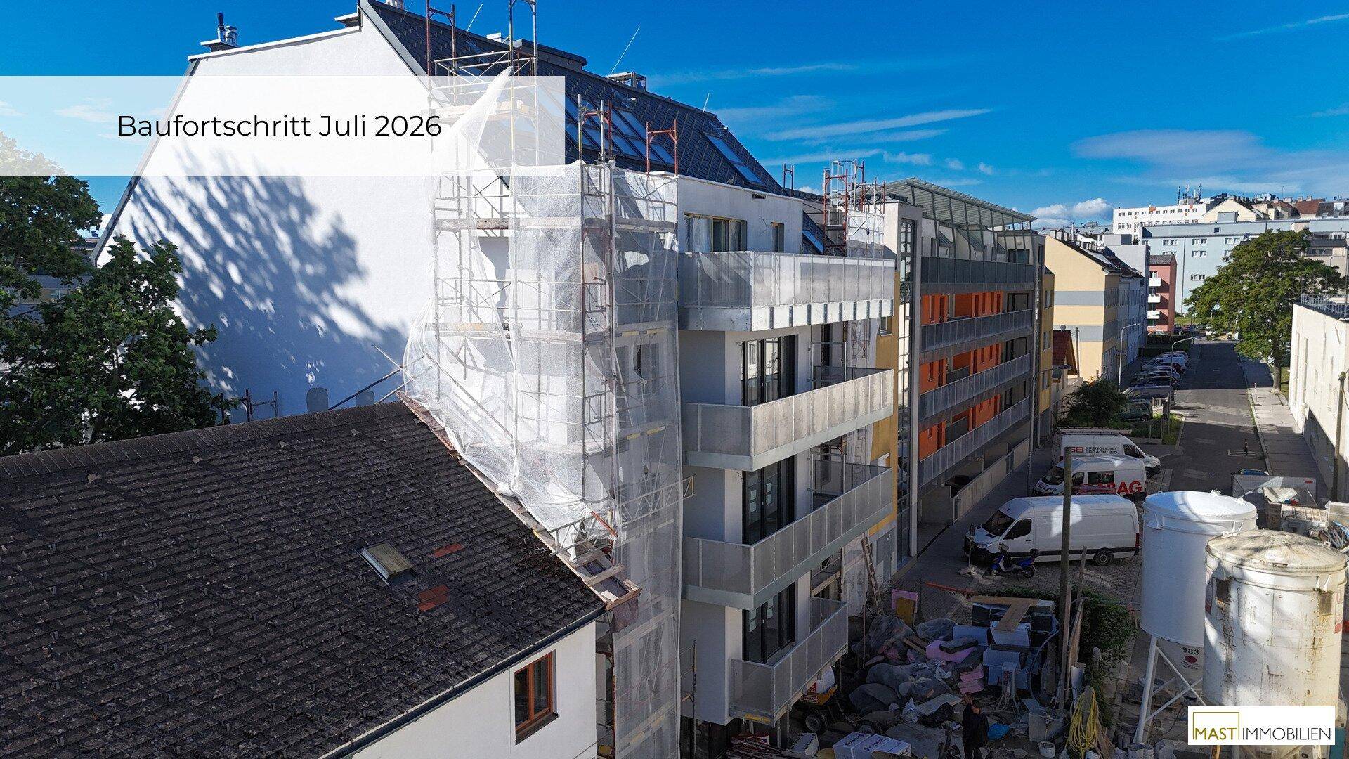 Luftaufnahme eines Neubaus im Bau mit Gerüst, modernen Balkonen und Mansarddach in städtischer Umgebung.