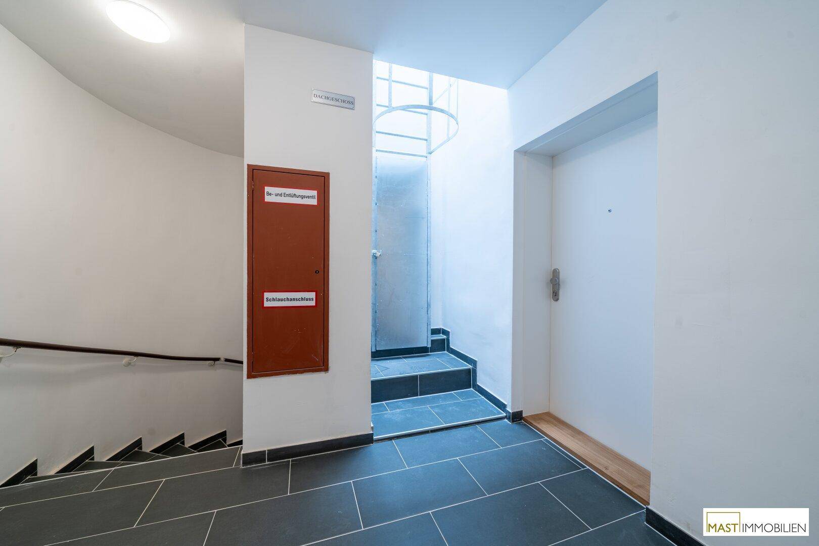 Heller Treppenhausbereich mit dunklen Fliesen, Treppe und Zugang zum Aufzug.