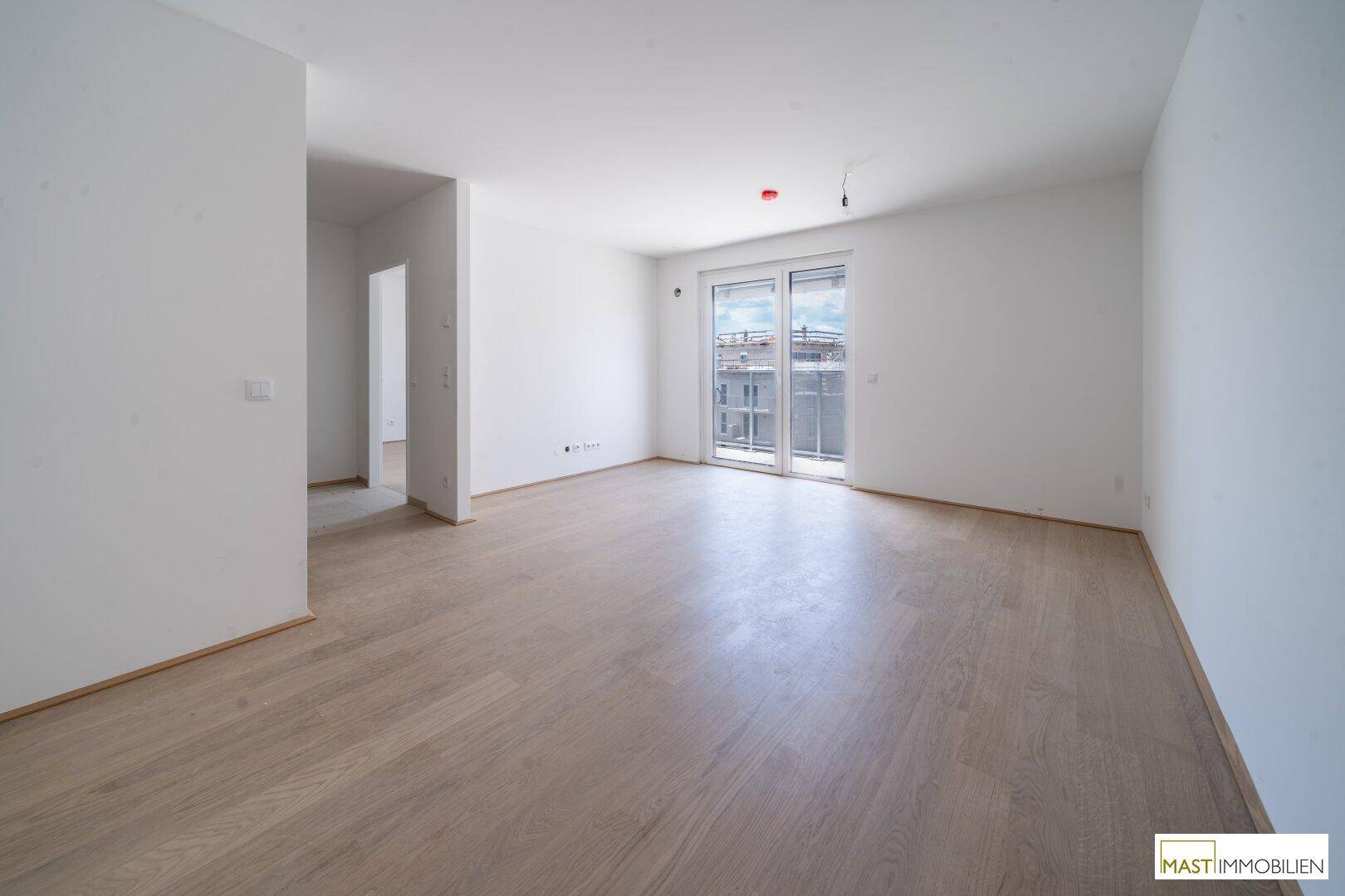 Heller, leerer Wohnraum mit hellem Holzfußboden, weißen Wänden und Zugang zu einem Balkon.