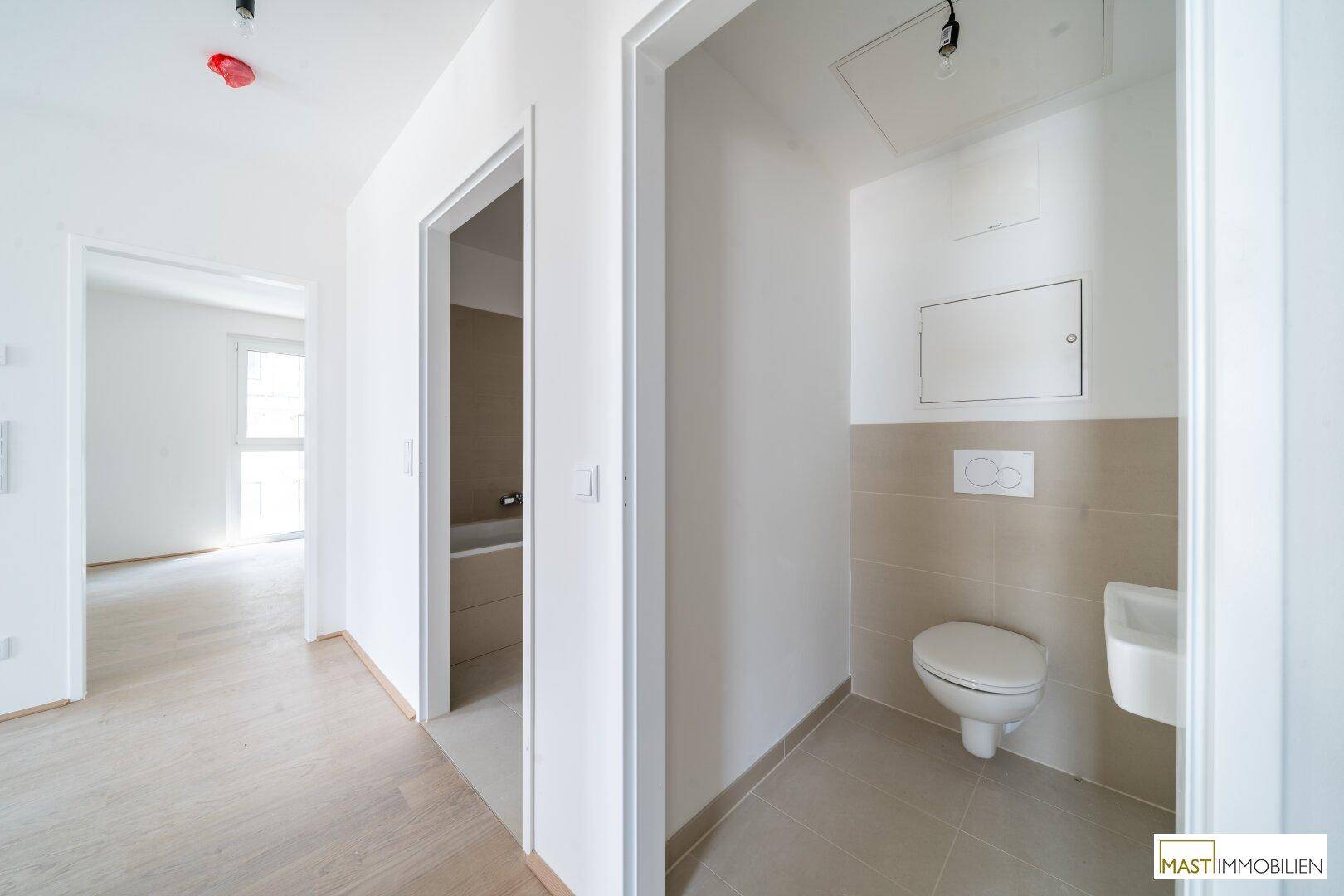 Separates WC mit wandmontierter Toilette, kleinem Waschbecken und beigefarbenen Wandfliesen.