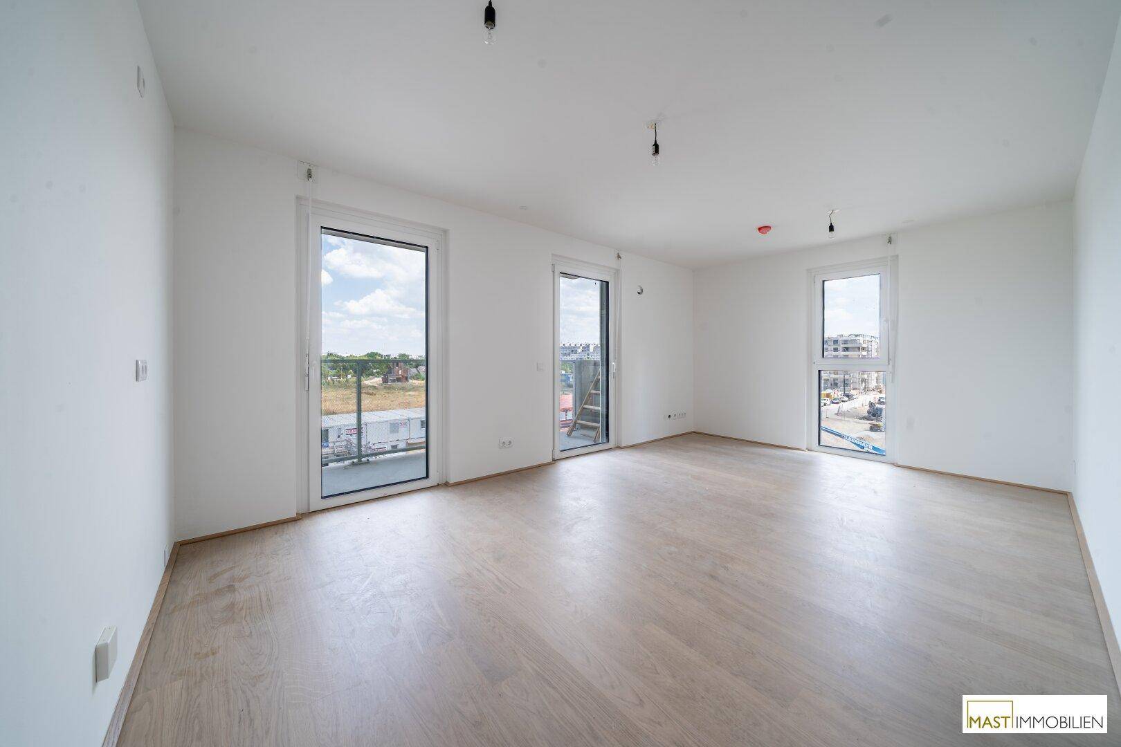 Helles, leeres Zimmer mit hellem Holzboden und mehreren Fenstern, die viel Tageslicht hereinlassen.