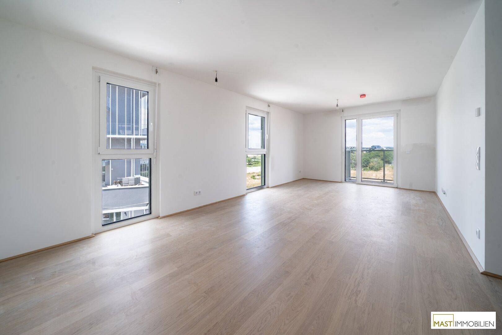 Helles, leeres Zimmer mit hellem Holzboden und mehreren Fenstern, die viel Tageslicht hereinlassen.