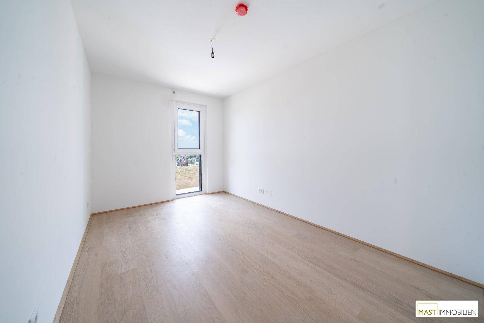 Leeres Schlafzimmer mit hellem Holzboden und einem großen Fenster, das den Raum erhellt.