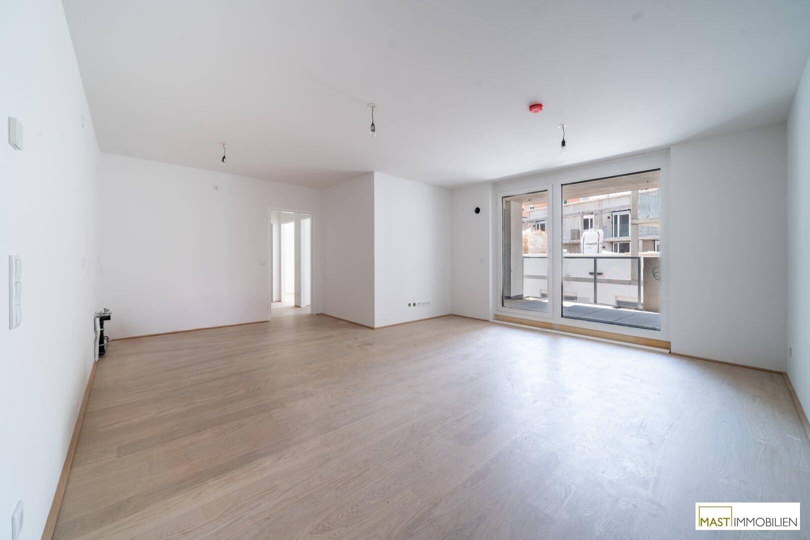 Helles, leeres Wohnzimmer mit hellem Holzboden und Zugang zu einem Balkon.