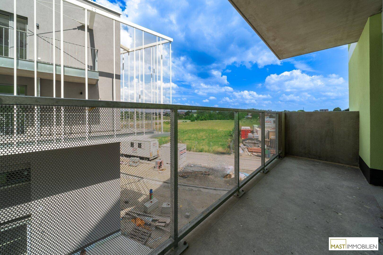 Balkon mit weitem Blick über eine grüne Wiese und eine Baustelle unter blauem Himmel.