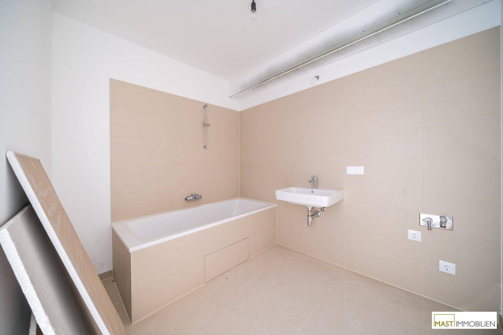 Unfertiges Badezimmer mit eingebauter Badewanne, Waschbecken und beigefarbenen Wandfliesen.