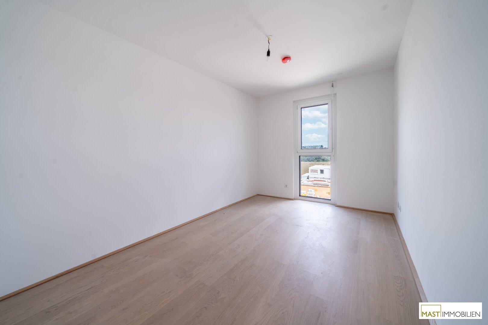Leeres Zimmer mit hellem Holzboden und einem Fenster, das viel Tageslicht hereinlässt.