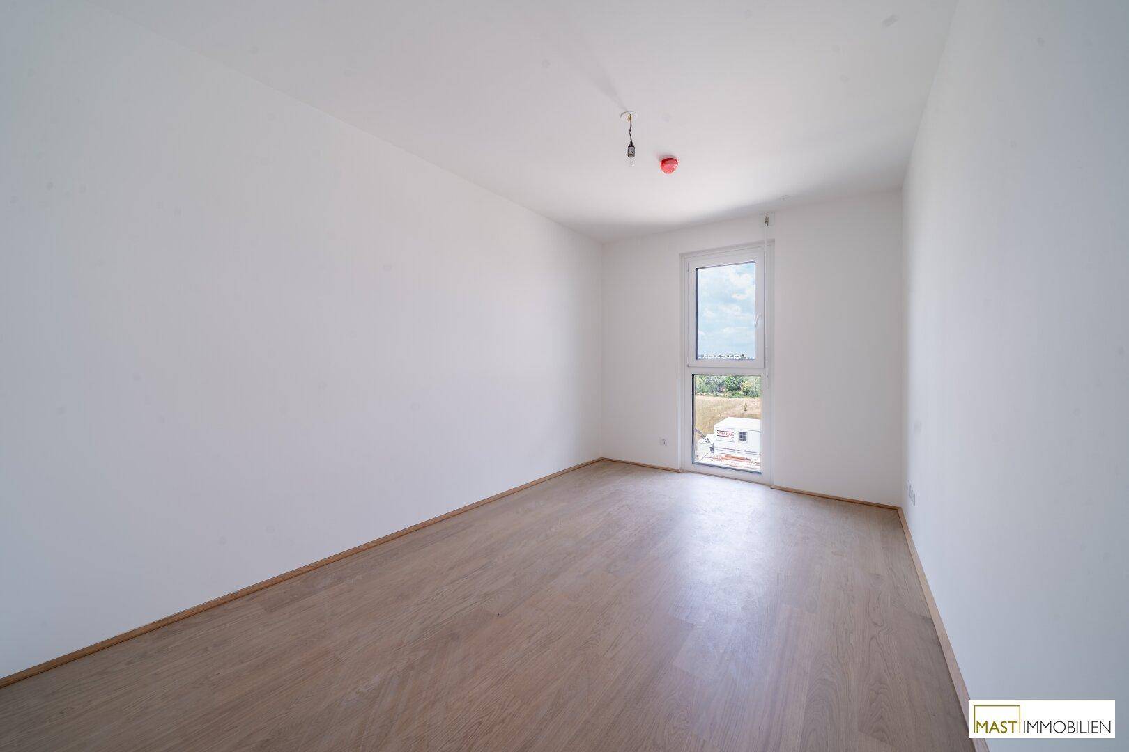 Helles Schlafzimmer mit Parkettboden und einem großen Fenster in einem modernen Neubau.