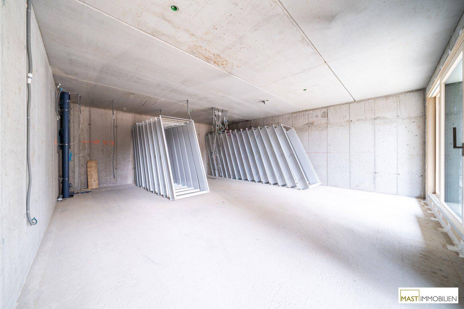 Rohbau-Innenraum mit Betonoberflächen, sichtbaren Installationen und vorbereiteten Fensterrahmen.