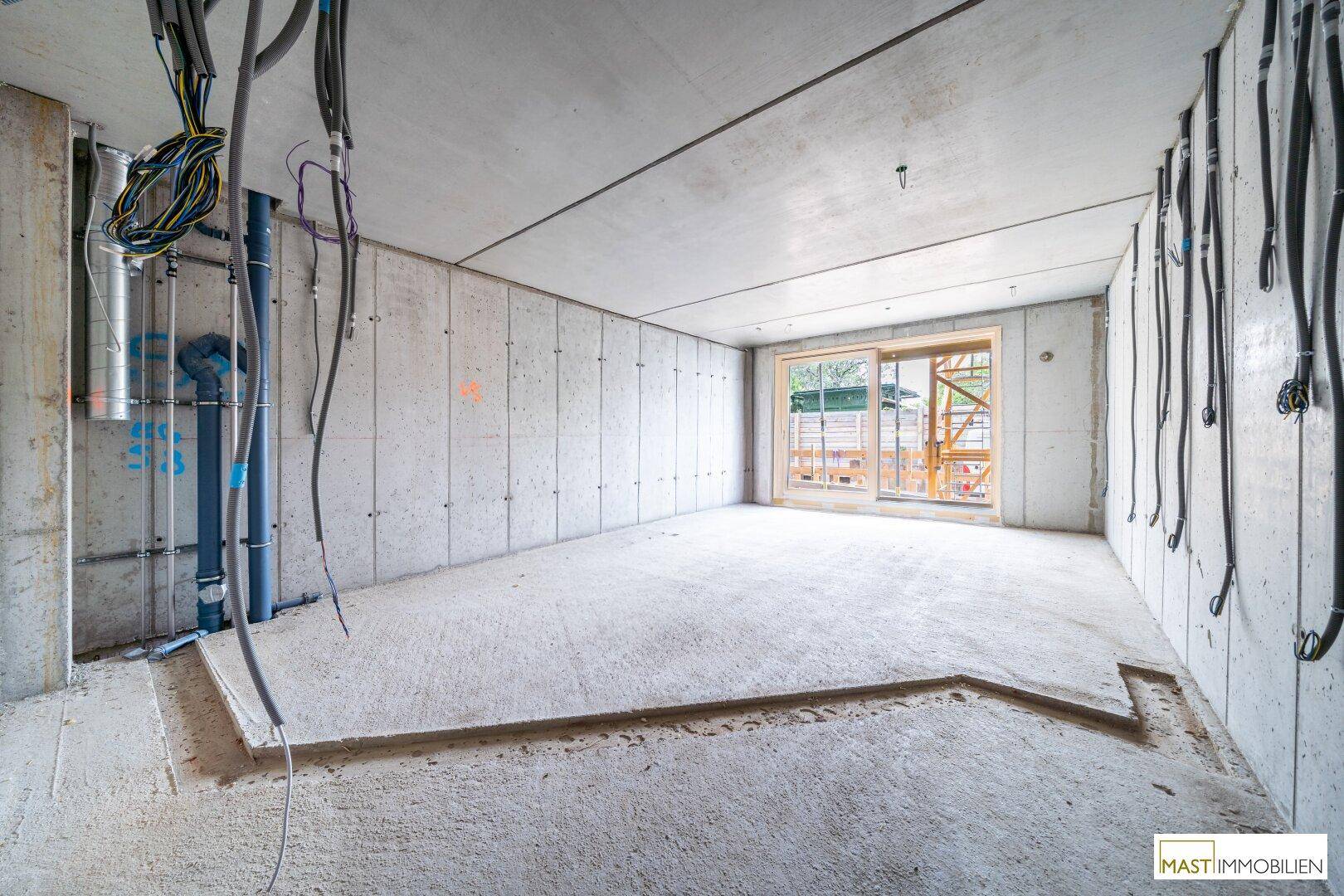 Rohbau-Innenraum mit Betonwänden, sichtbaren Installationen und großem Fenster zur Baustelle.