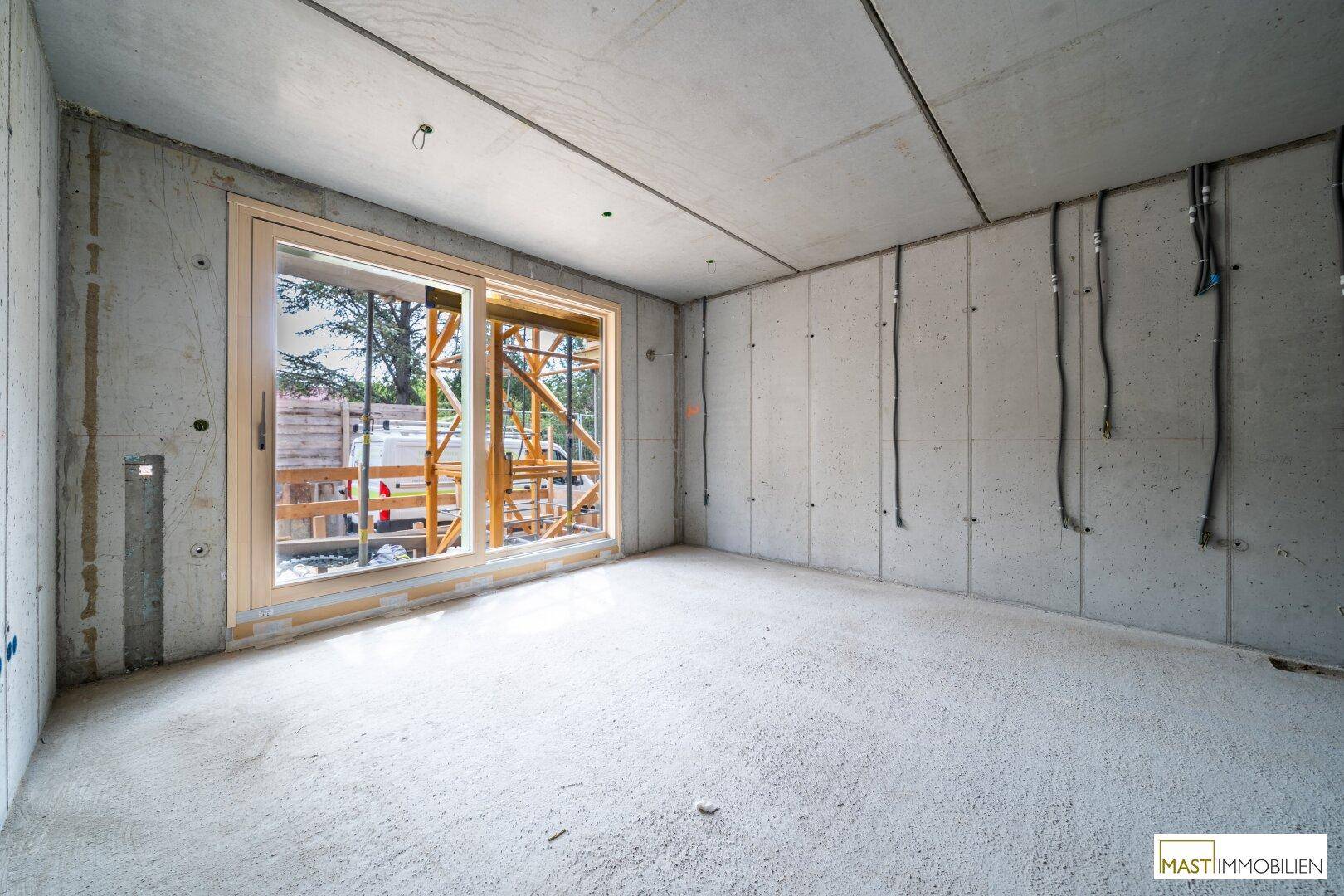 Heller Rohbau-Innenraum mit Betonwänden, sichtbaren Leitungen und großem Fenster zur Baustelle.