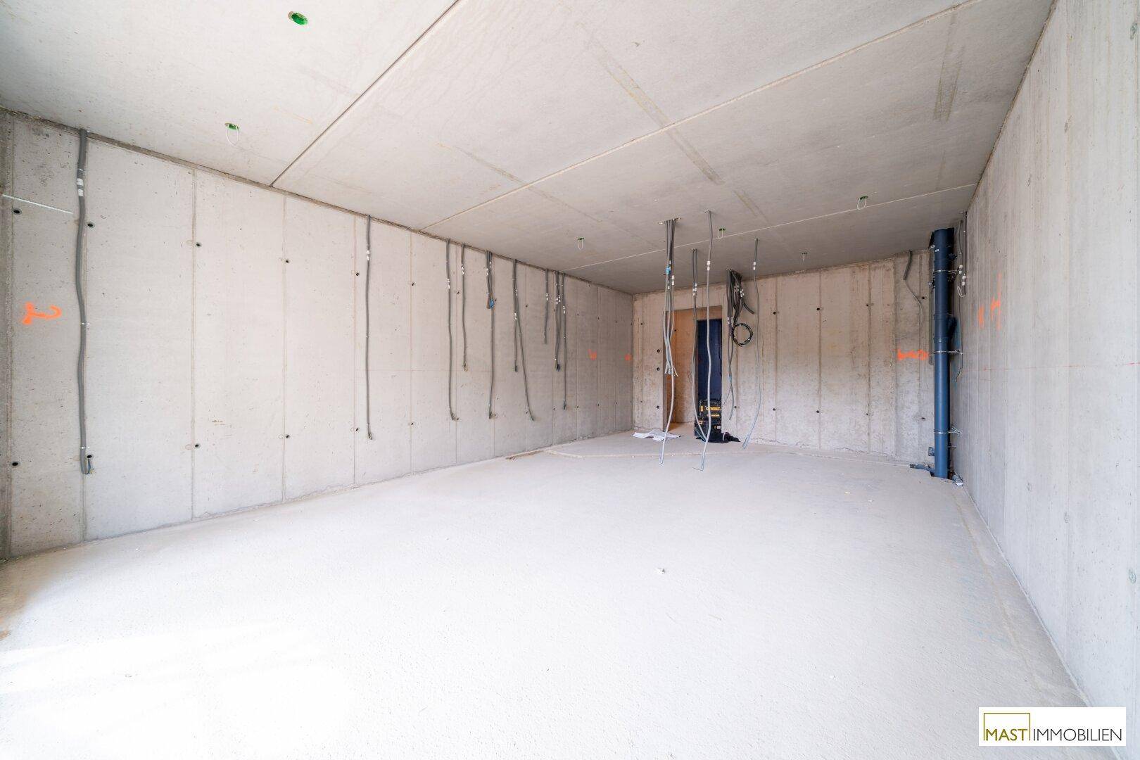 Unfertiger Innenraum mit Betonwänden, sichtbaren Kabeln und Rohren im Rohbauzustand.