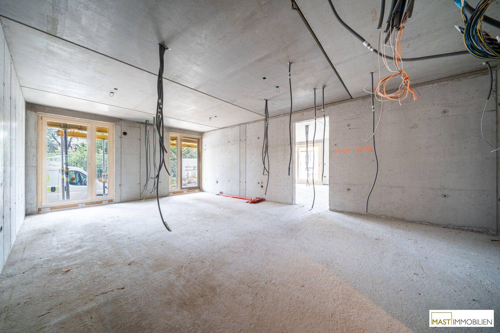 Unfertiger Innenraum eines Neubaus mit Betonwänden, sichtbaren Kabeln und großen Fensteröffnungen.
