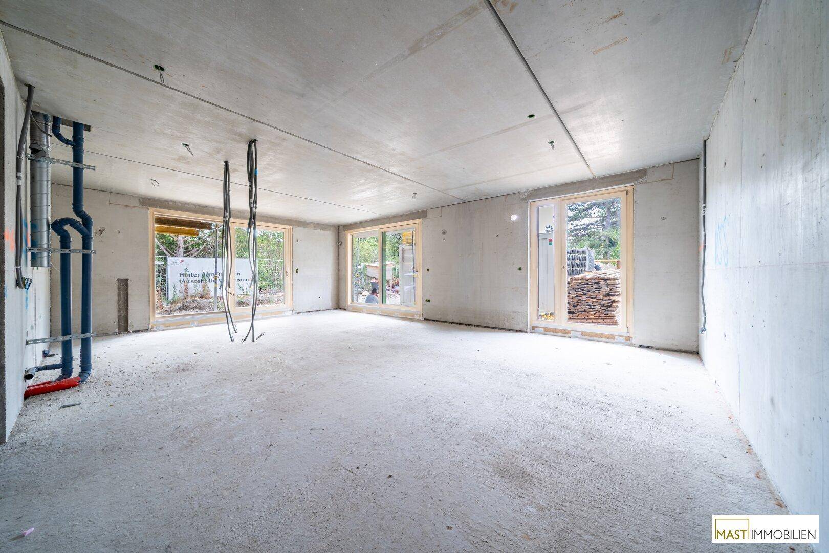 Heller Rohbau-Raum mit Betonoberflächen, sichtbaren Leitungen und Zugang zu Außenbereichen.