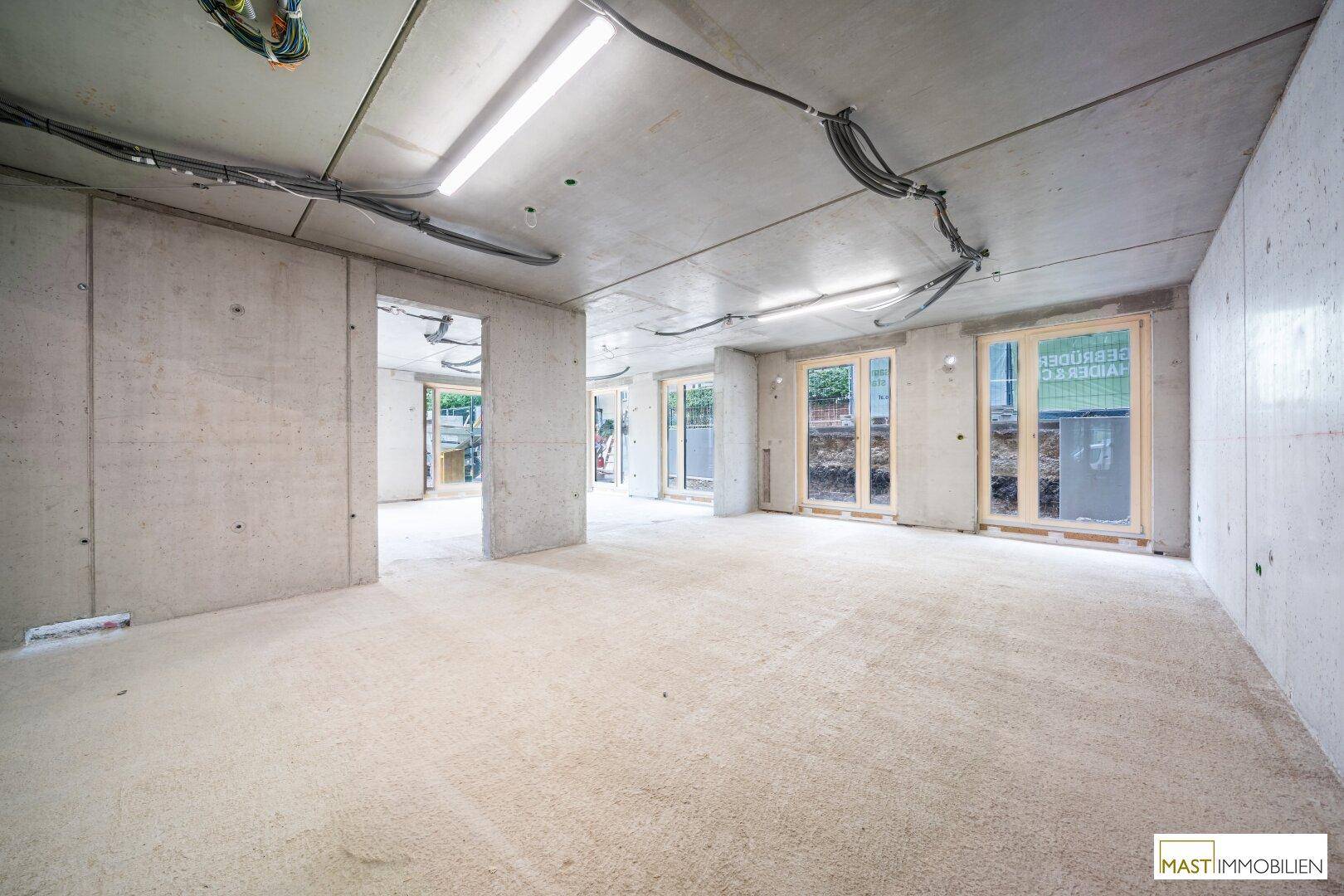Rohbau-Innenraum mit Betonoberflächen, sichtbaren Leitungen und mehreren Fensteröffnungen.