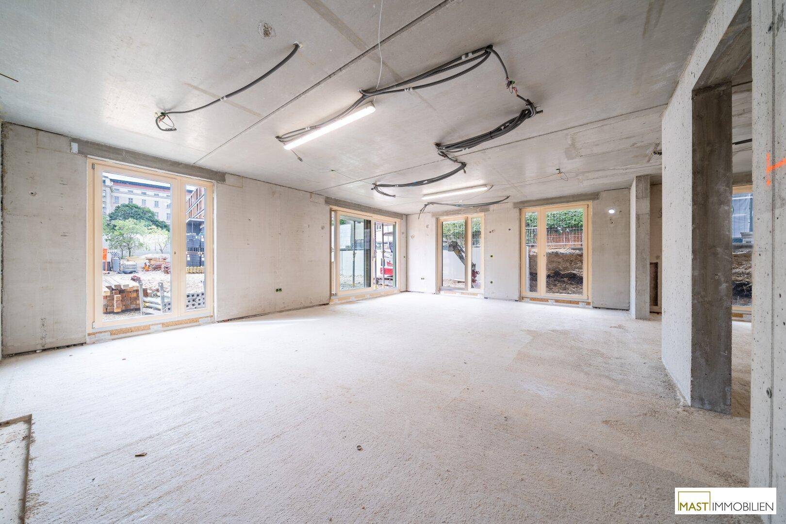 Geräumiger Rohbau-Innenraum mit Betonwänden, Decken und großen Fensterfronten zum Außenbereich.