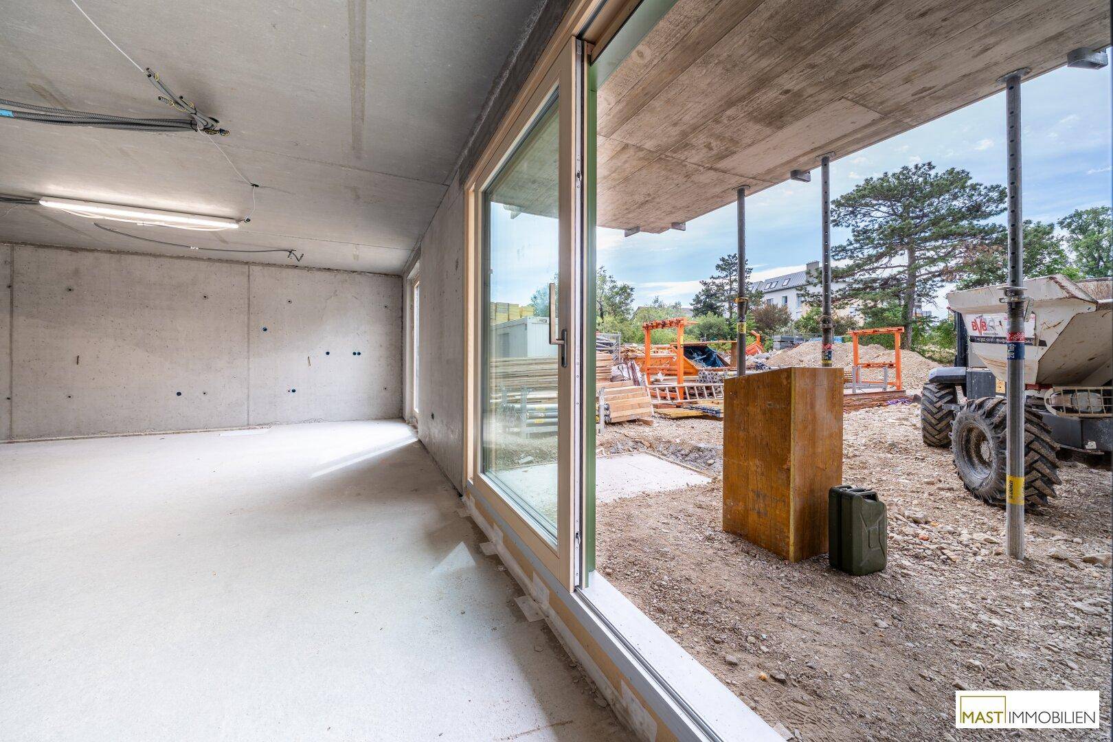 Blick aus einem Rohbau durch eine große Fensteröffnung auf eine Baustelle im Freien.