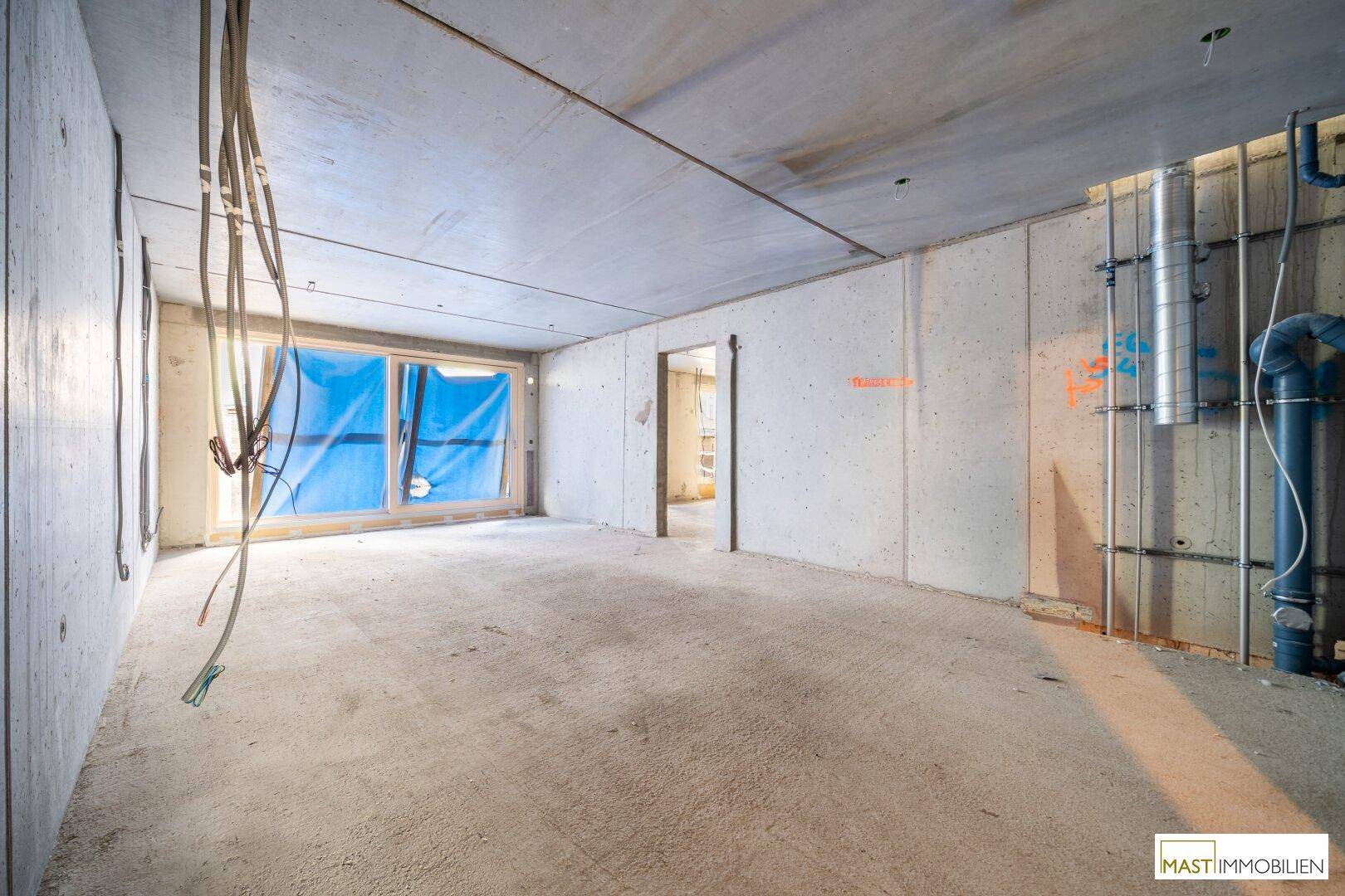 Unfertiger Innenraum im Rohbauzustand mit sichtbaren Installationen und Fenstern, bereit für den Ausbau.