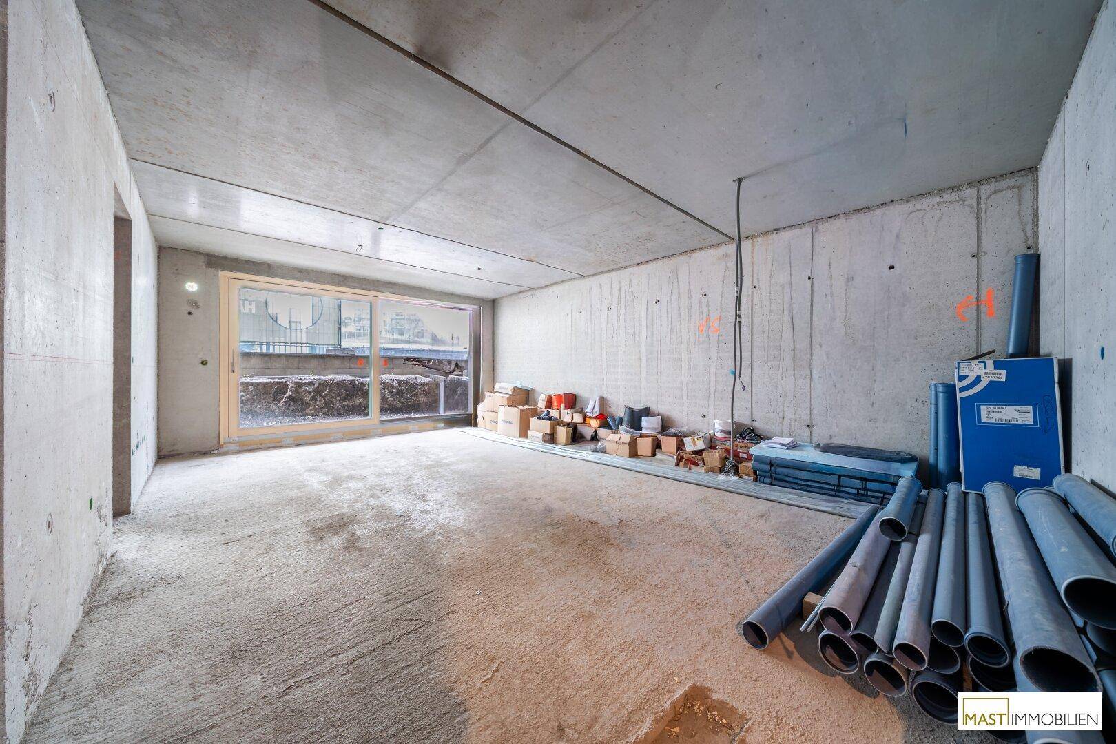 Unfertiger Innenraum mit Betonwänden und -boden, großen Fensteröffnungen und Baumaterialien.