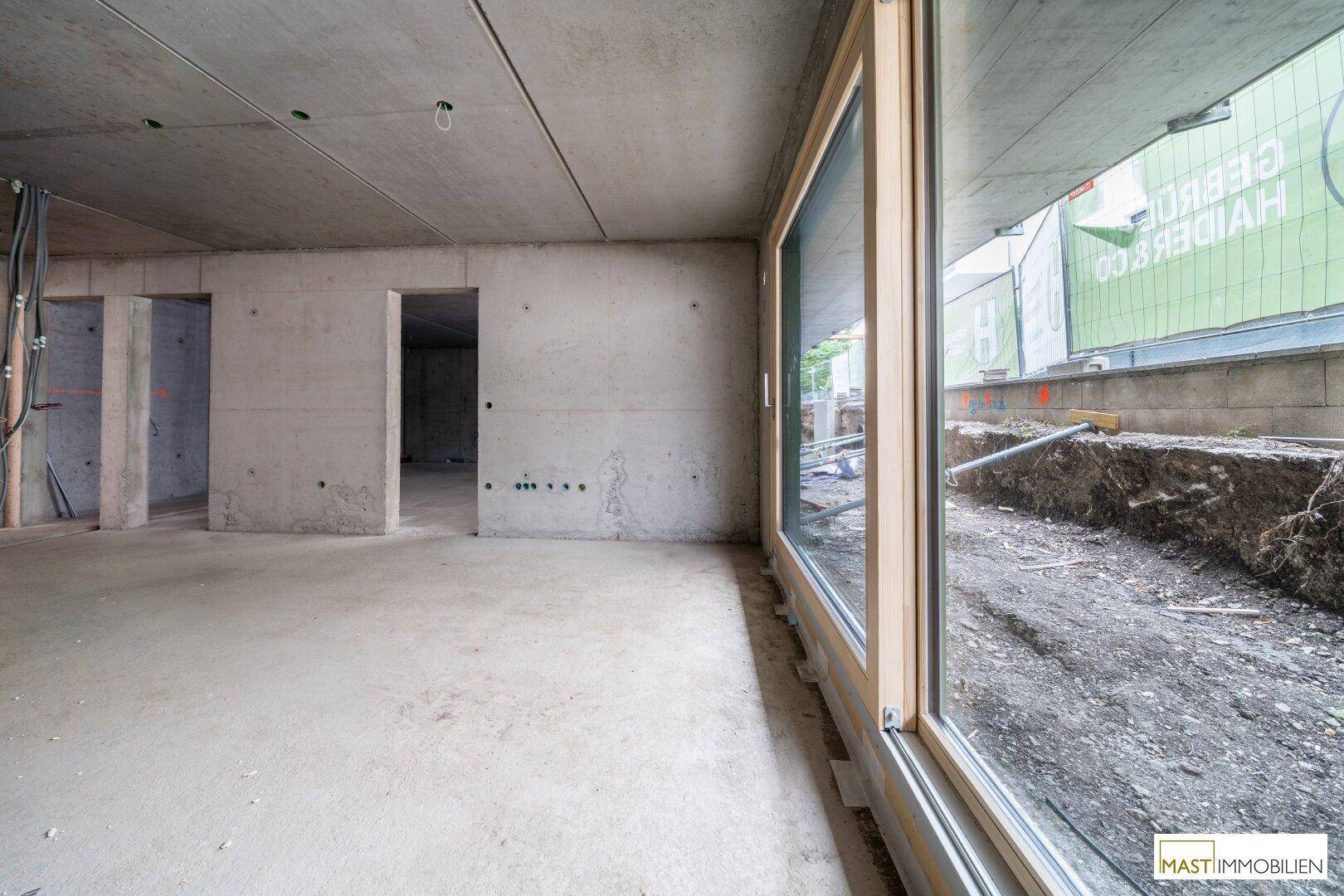 Unfertiger Raum mit großen Fenstern, Betonwänden und Blick auf die Baustelle im Außenbereich.