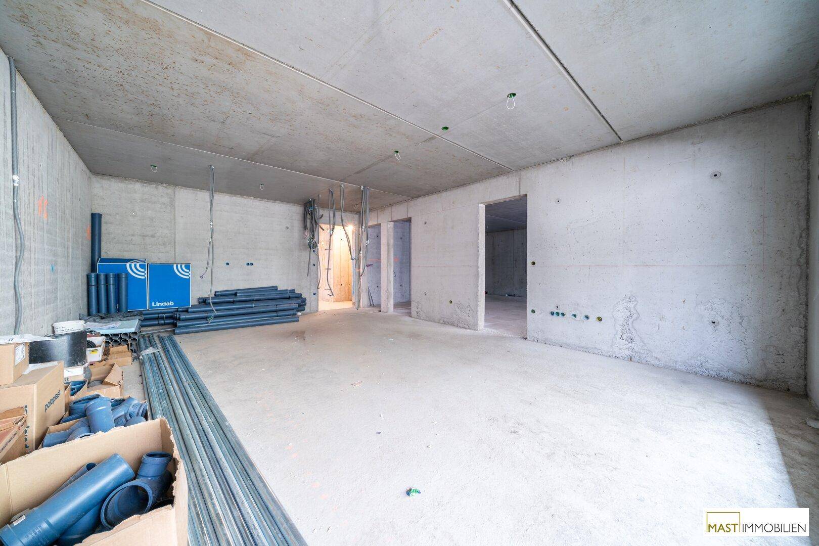 Rohbau-Innenraum mit sichtbaren Betonstrukturen, mehreren Türöffnungen und Baumaterialien.