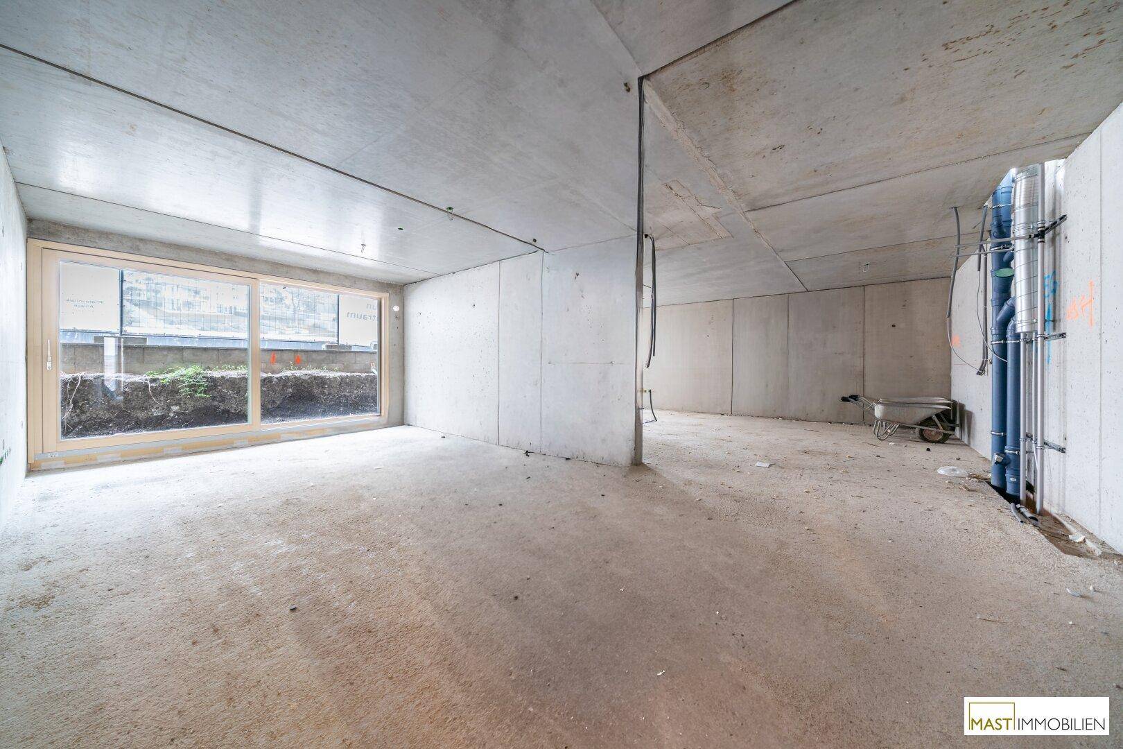 Unfertiger Innenraum mit Betonwänden und großem Fenster, das Tageslicht hereinlässt.