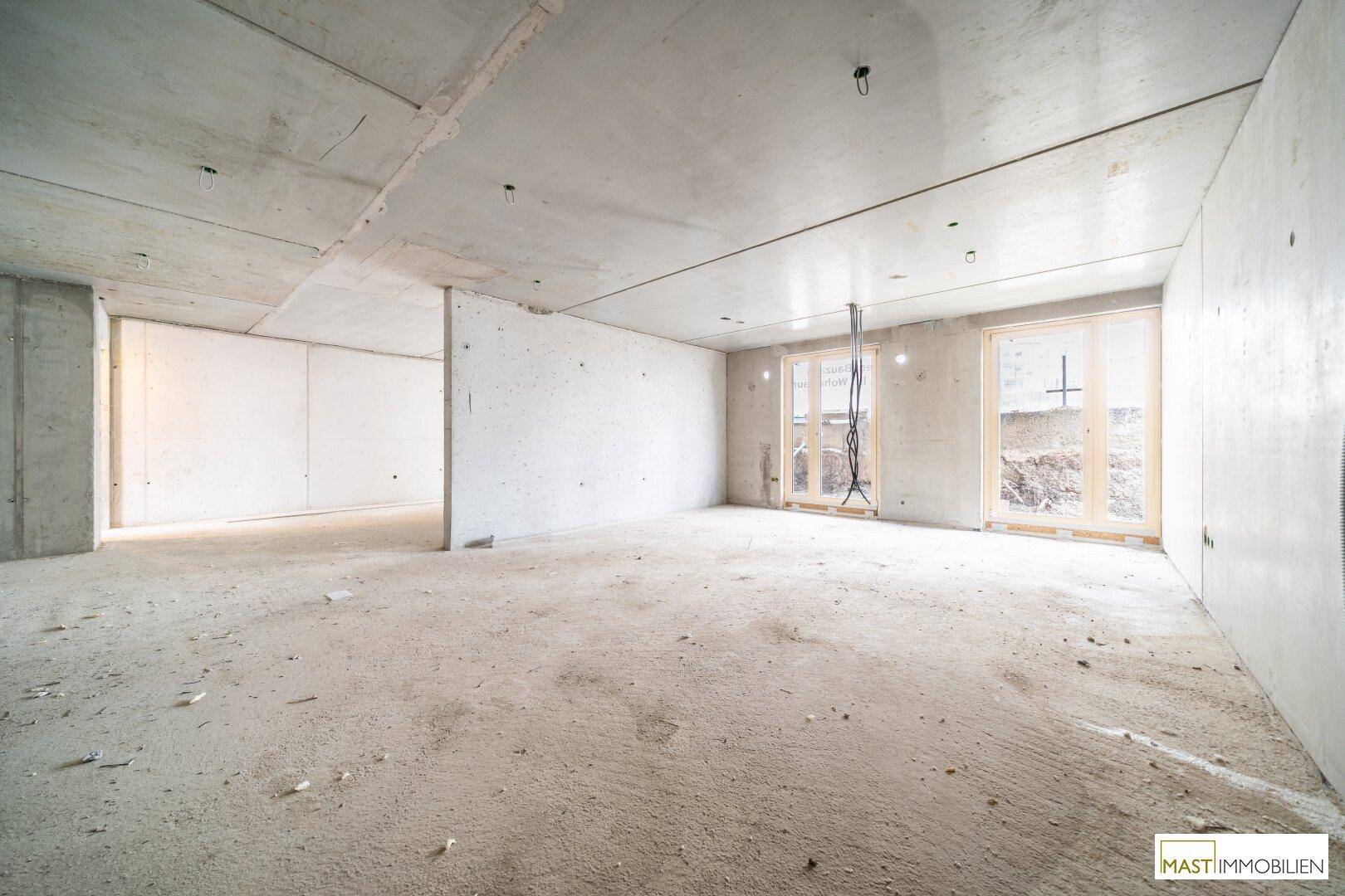 Rohbau-Innenraum mit Betonoberflächen und zwei Fensteröffnungen, die auf den Außenbereich blicken.
