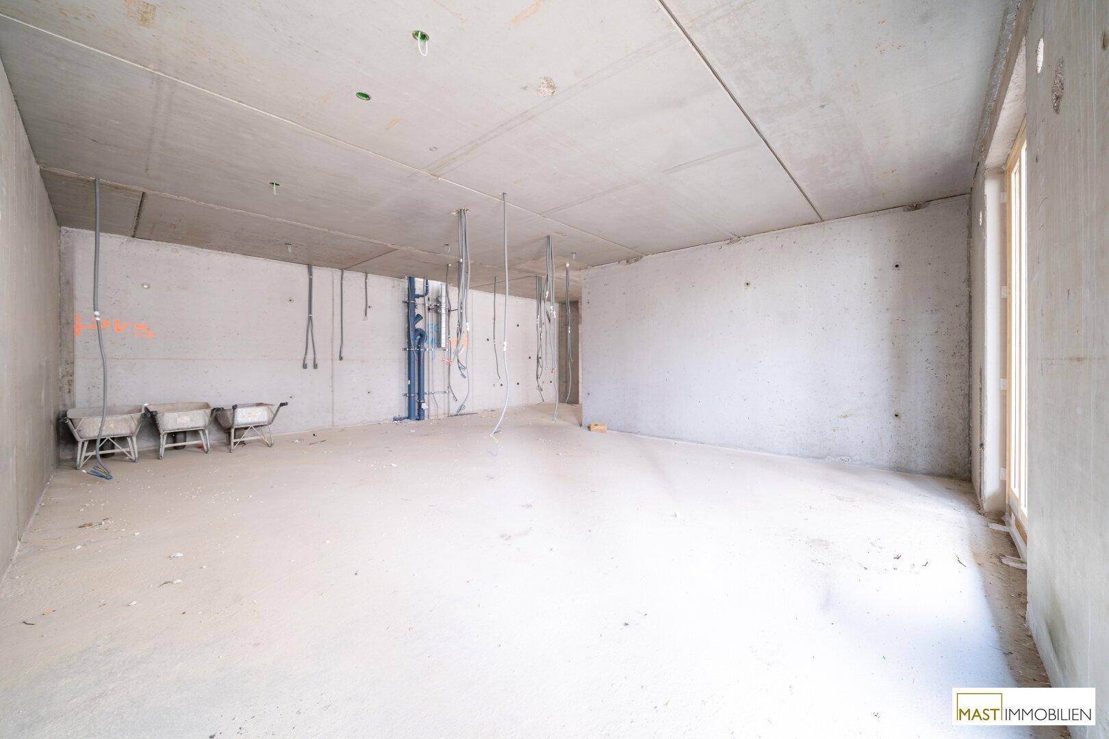 Leerer Rohbau mit Betonwänden, sichtbaren Installationen und einer Fensteröffnung für natürliches Licht.