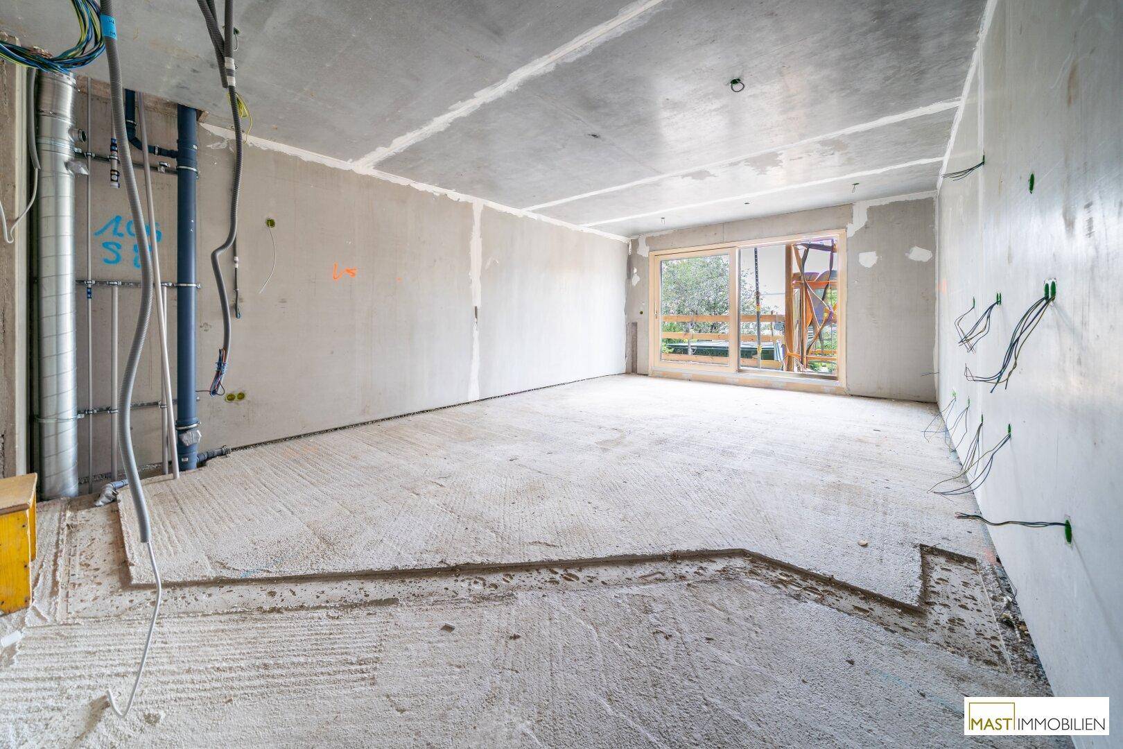 Rohbau eines Raumes mit Betonwänden und -boden sowie sichtbaren Installationen und großem Fenster.