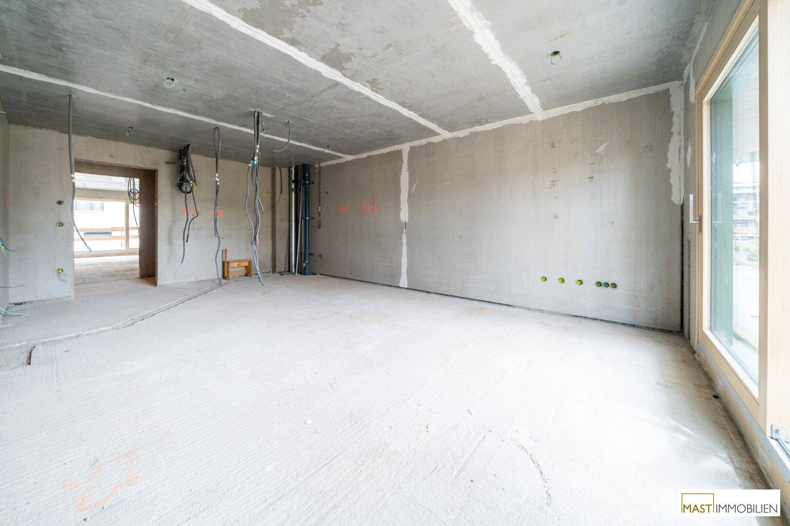 Unfertiger Innenraum mit Betonoberflächen, vorbereiteten Elektroinstallationen und Zugang zu weiteren Räumen.