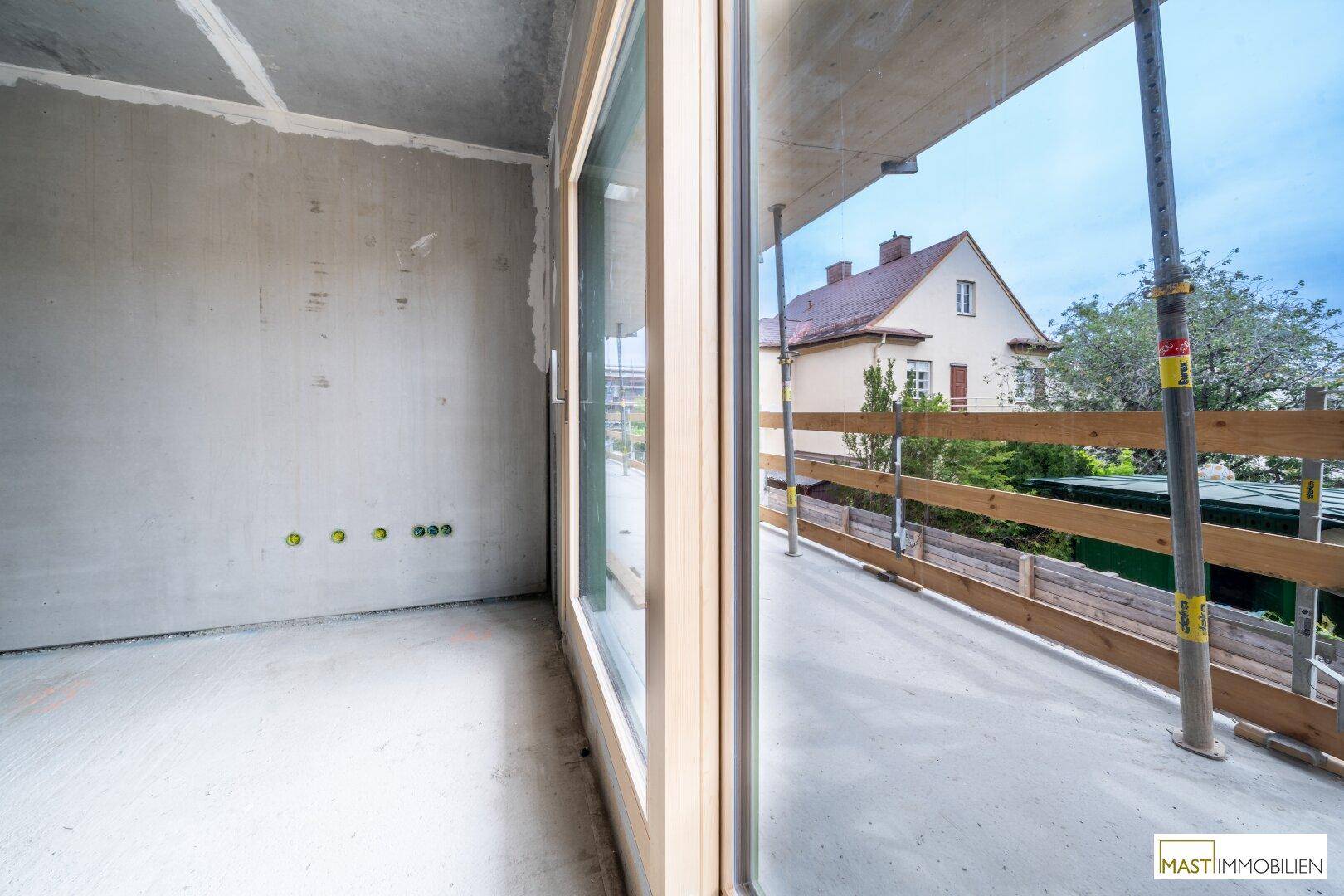 Blick aus einem Rohbauraum durch ein großes Fenster auf die Baustelle und ein benachbartes Gebäude.