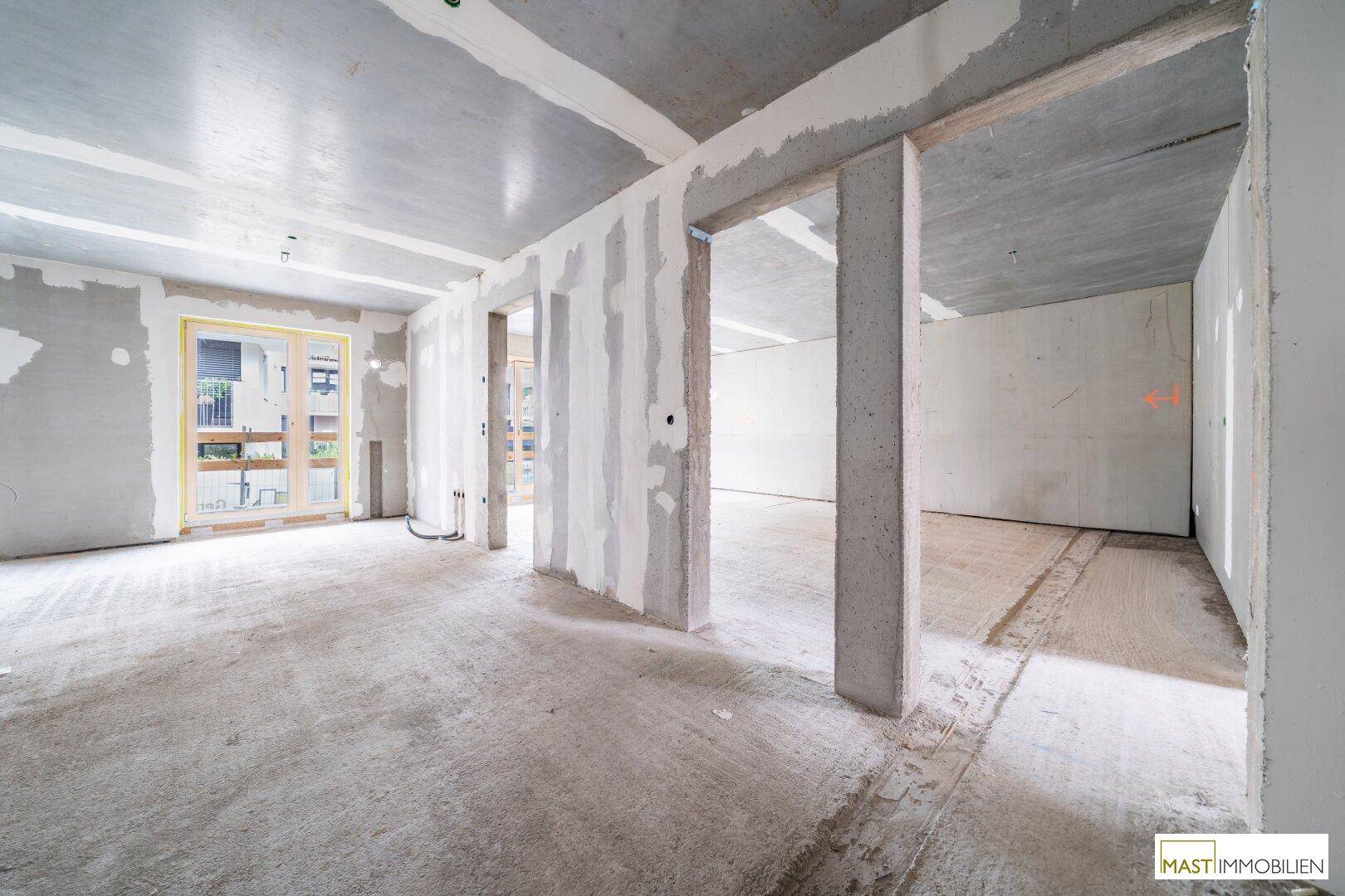 Rohbau-Innenraum mit großen Fenstern und Zugang zu einem zukünftigen Außenbereich.