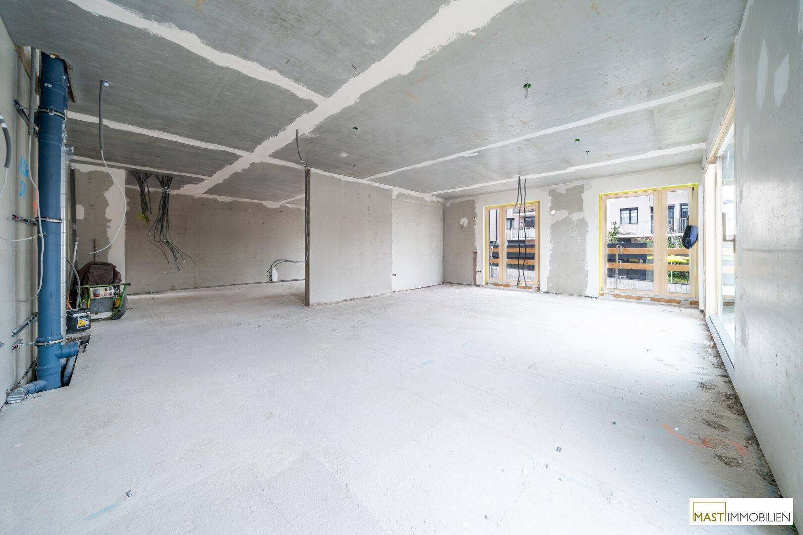 Geräumiger Rohbau-Innenraum mit sichtbaren Leitungen und Fenstern, die den Blick nach draußen freigeben.