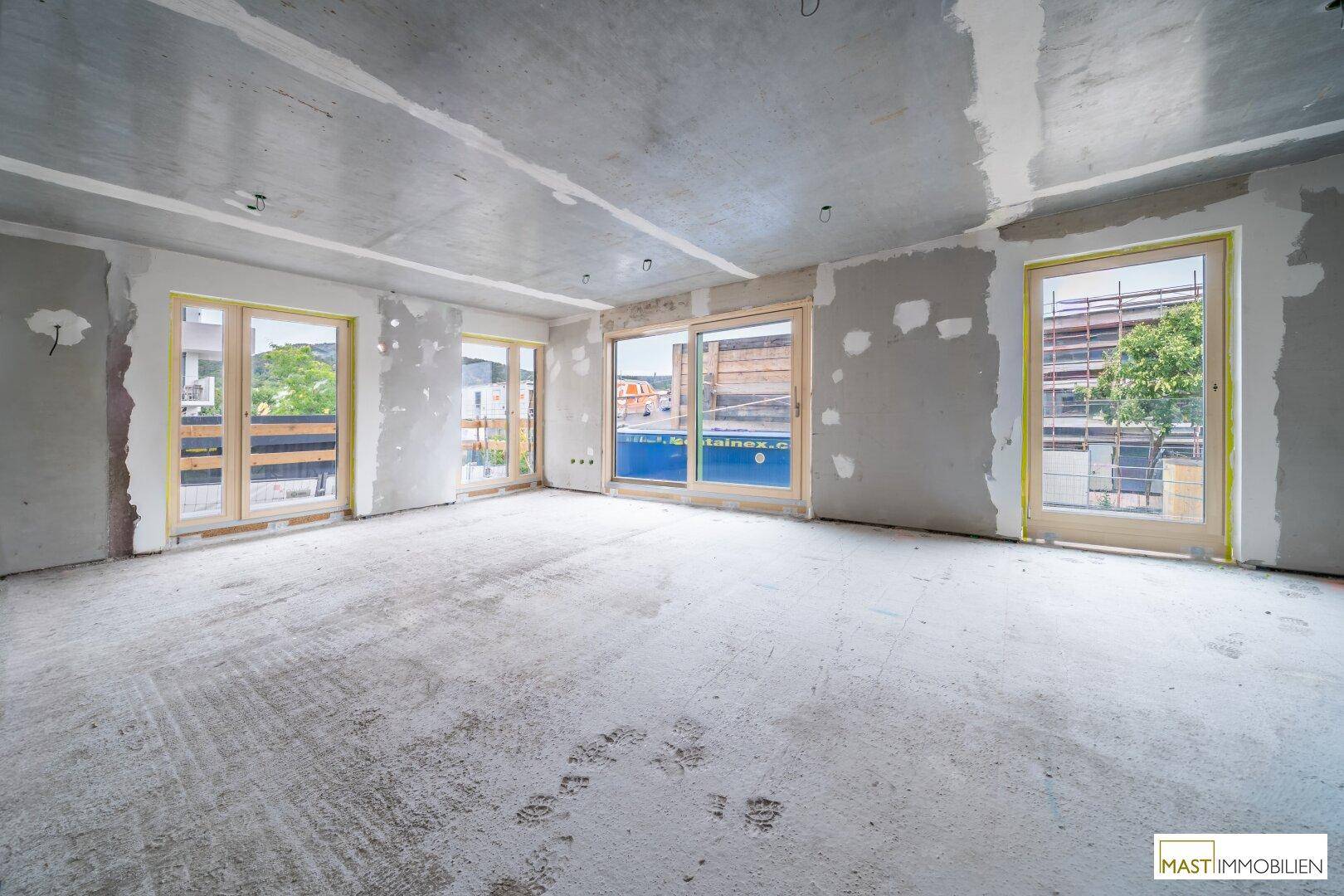 Großer, heller Rohbau-Raum mit mehreren Fenstern, die eine weite Aussicht bieten.