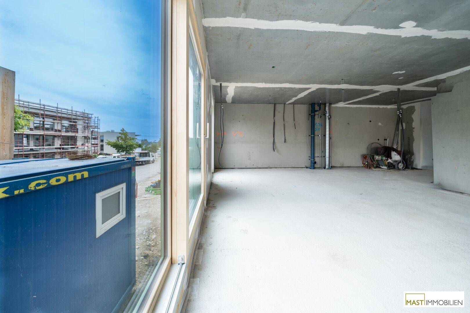 Innenraum im Rohbau mit großem Fenster, das einen Blick auf die Baustelle und den blauen Himmel ermöglicht.
