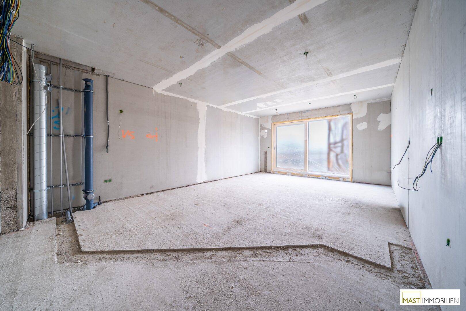 Unfertiger Innenraum mit Betonwänden, sichtbaren Installationen und großem Fenster, bereit für den Ausbau.