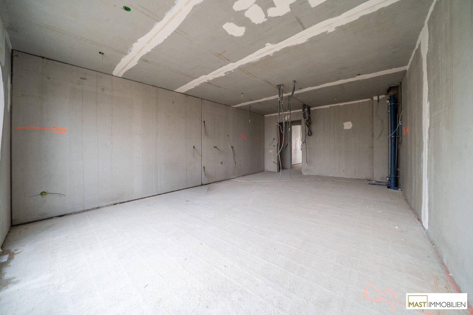 Rohbau eines Innenraums mit Betonwänden und -boden, sichtbaren Leitungen und Türöffnung.