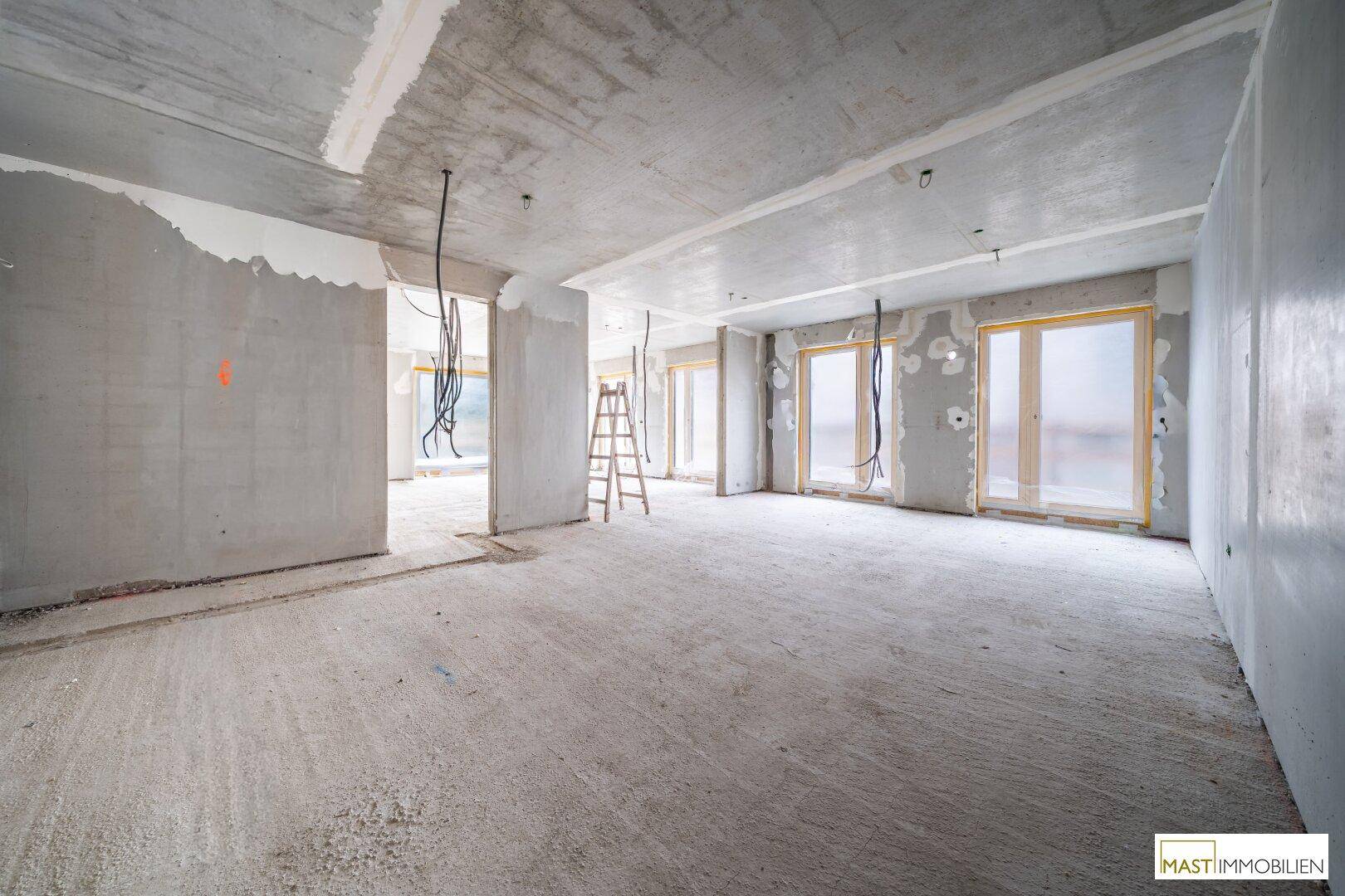 Unfertiger Innenraum mit Betonwänden und -boden, großen Fenstern und sichtbaren Installationen.