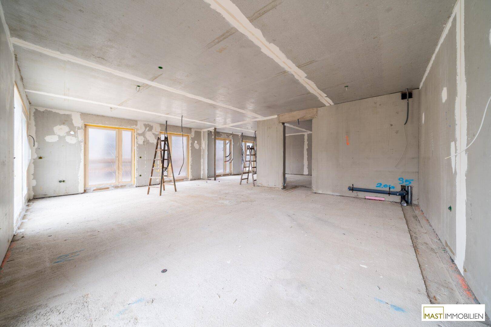 Unfertiger Raum mit sichtbaren Rohrleitungen, Betonwänden und -boden sowie großen Fensteröffnungen.