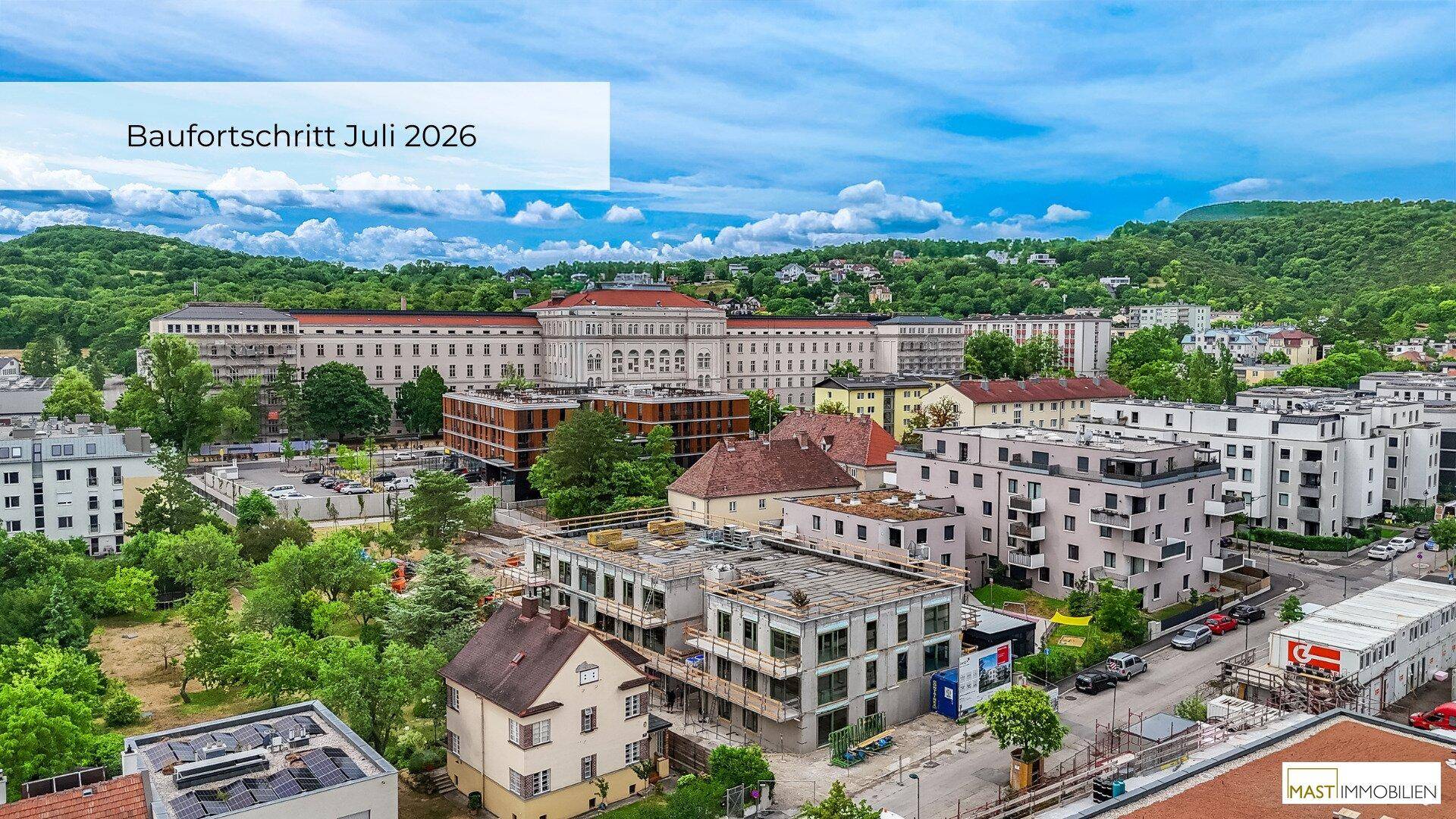 Luftaufnahme einer Baustelle mit umliegenden Gebäuden und grüner Landschaft im Hintergrund.