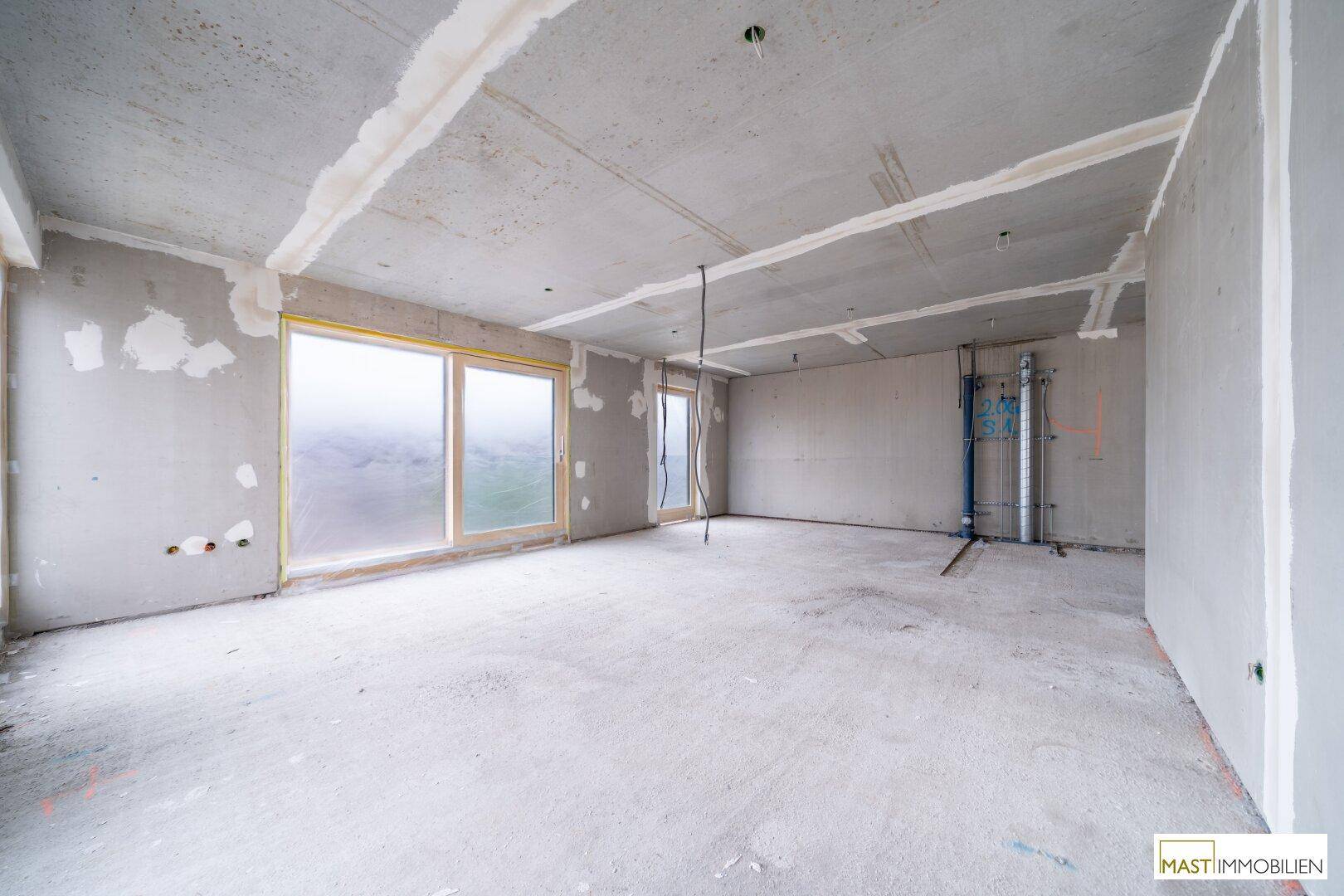 Rohbau eines Raumes mit Betonboden, großen Fenstern und sichtbaren Installationen an der Wand.