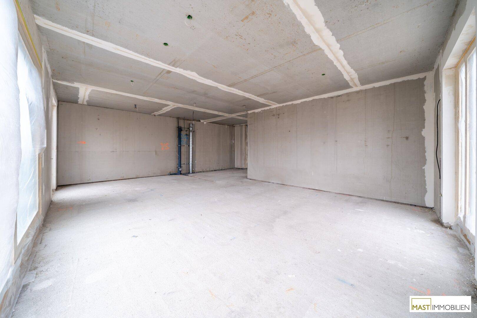 Unfertiger Innenraum mit Betonboden und einer Fensterfront, bereit für den weiteren Ausbau.