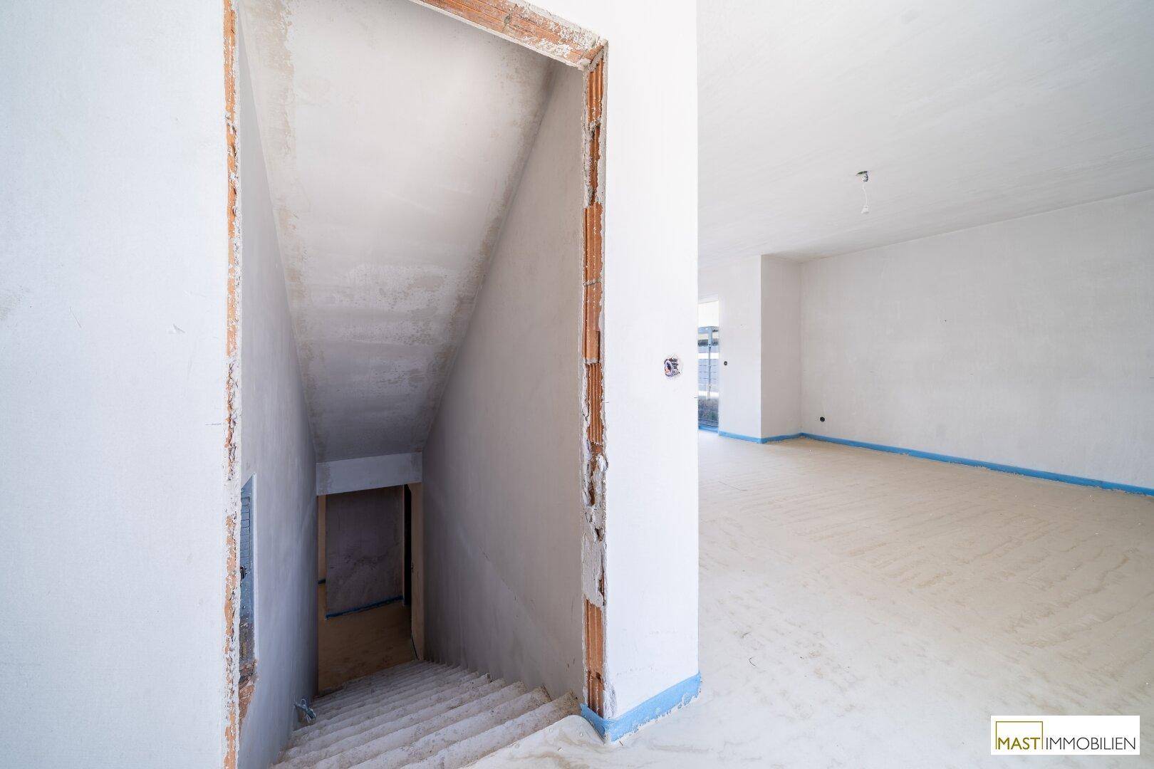 Unfertiger Innenraum mit Treppenabgang und weiß verputzten Wänden, bereit für den weiteren Ausbau.
