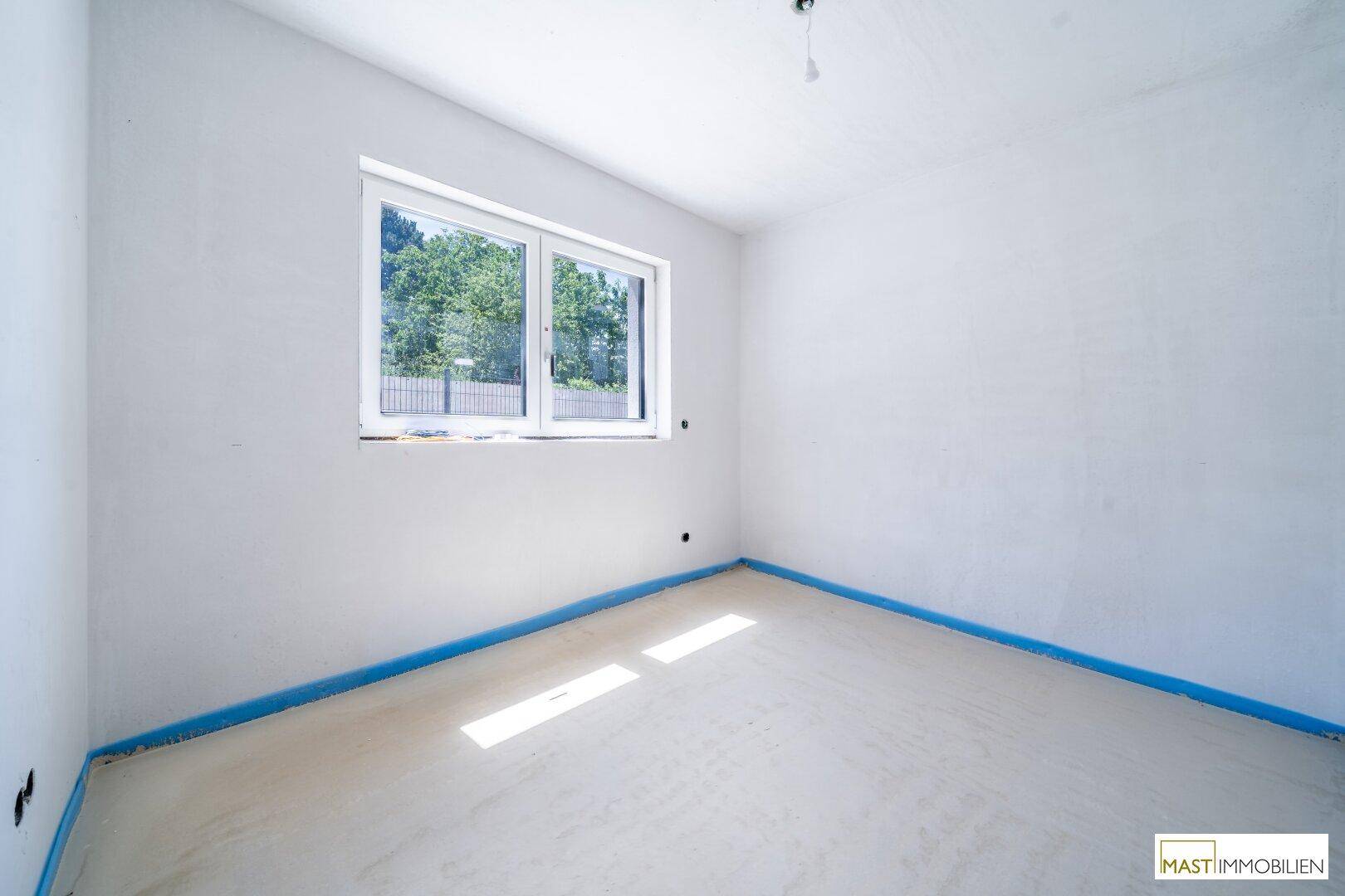 Unfertiges Schlafzimmer mit einem großen Fenster, das viel Tageslicht hereinlässt und einen grünen Ausblick bietet.